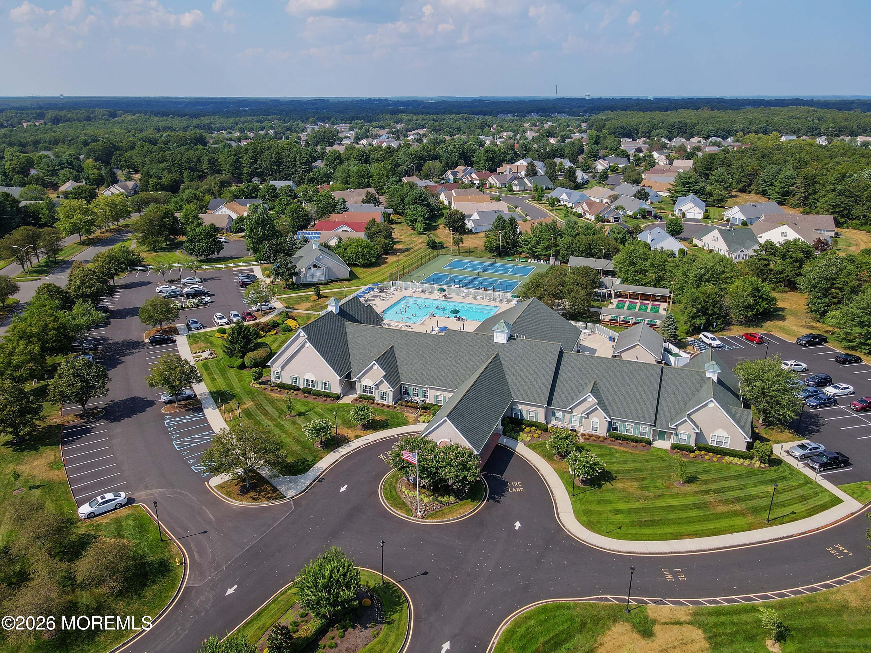 Aerial view of a residential community clubhouse, pool, and tennis courts surrounded by homes and trees.