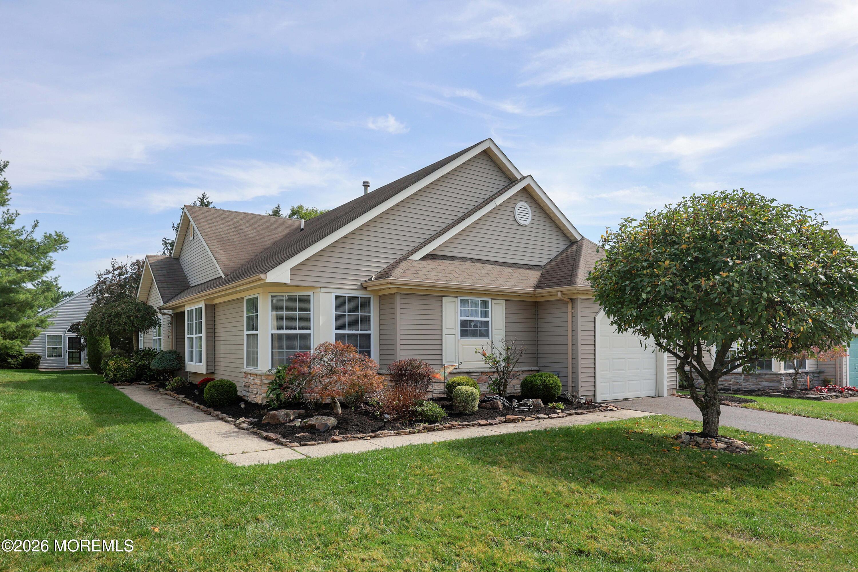 Beige-sided home with gabled rooflines, attached garage, landscaped shrubs, and a mature front-yard tree.
