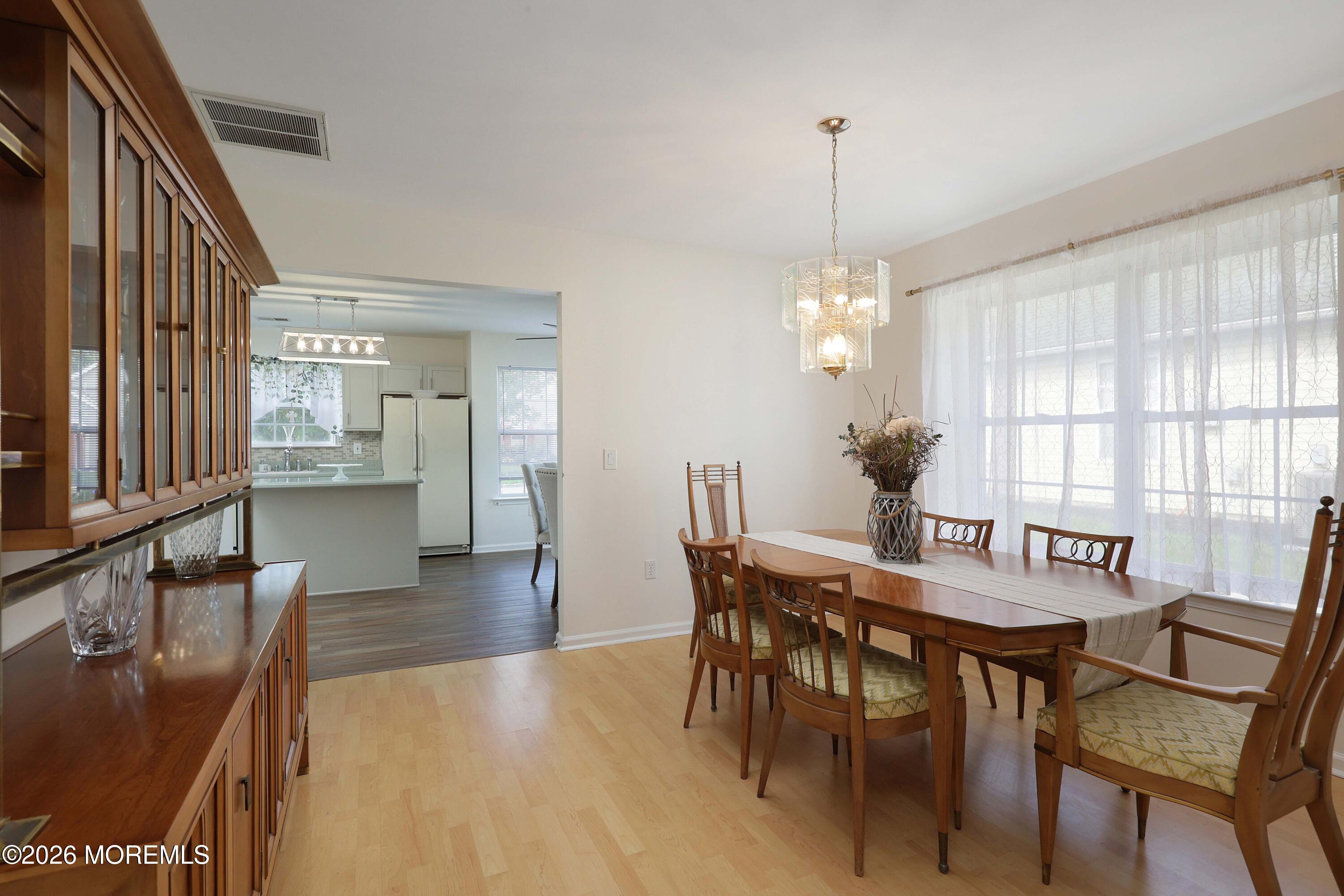 Dining room with a wooden table beside large windows, opening to a kitchen with a breakfast counter.