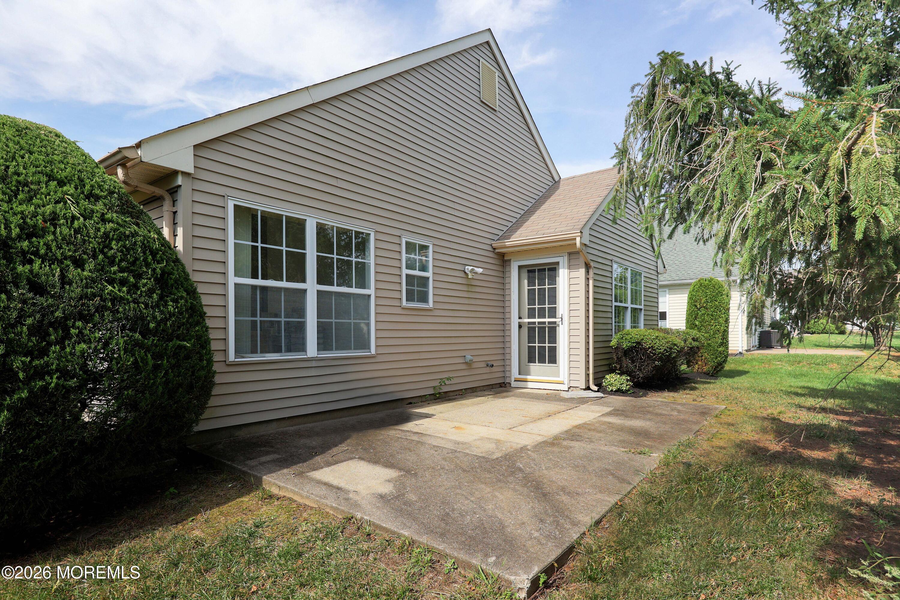 Rear of a tan-sided house with a concrete patio, screened door, and mature landscaping.