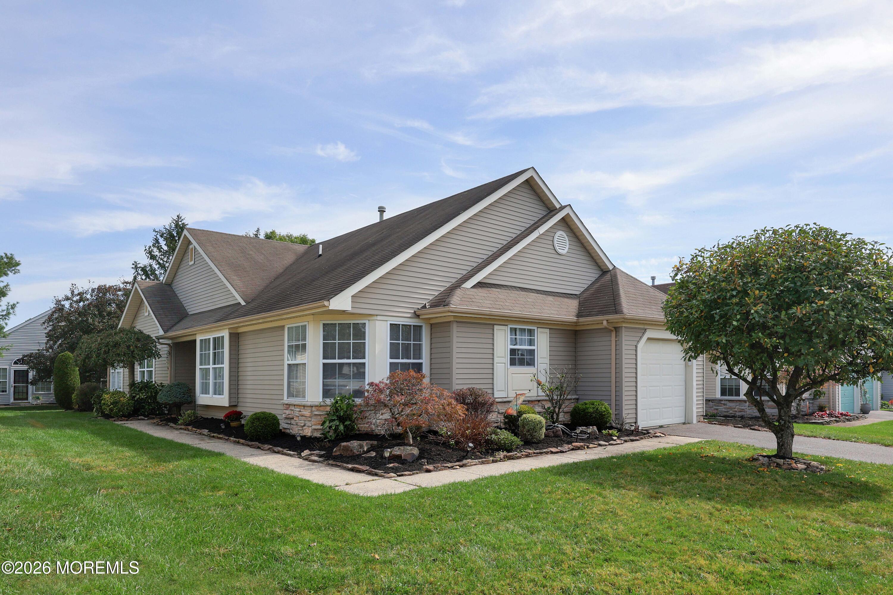 Beige multi-gabled house with a garage, landscaped foundation beds, and a mature front-yard tree.