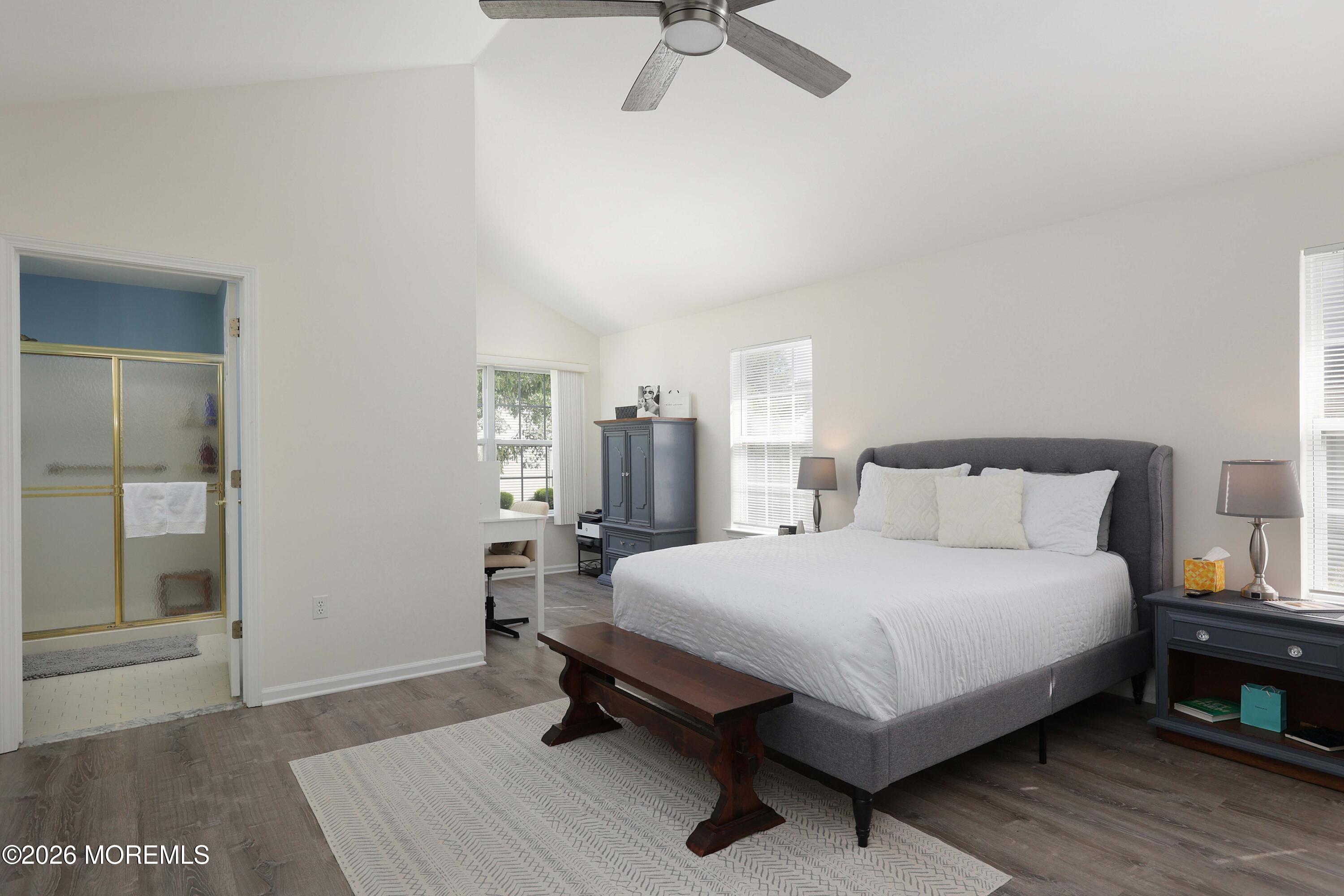 Furnished bedroom with a gray upholstered bed, white linens, dark wood bench, and adjoining bathroom.