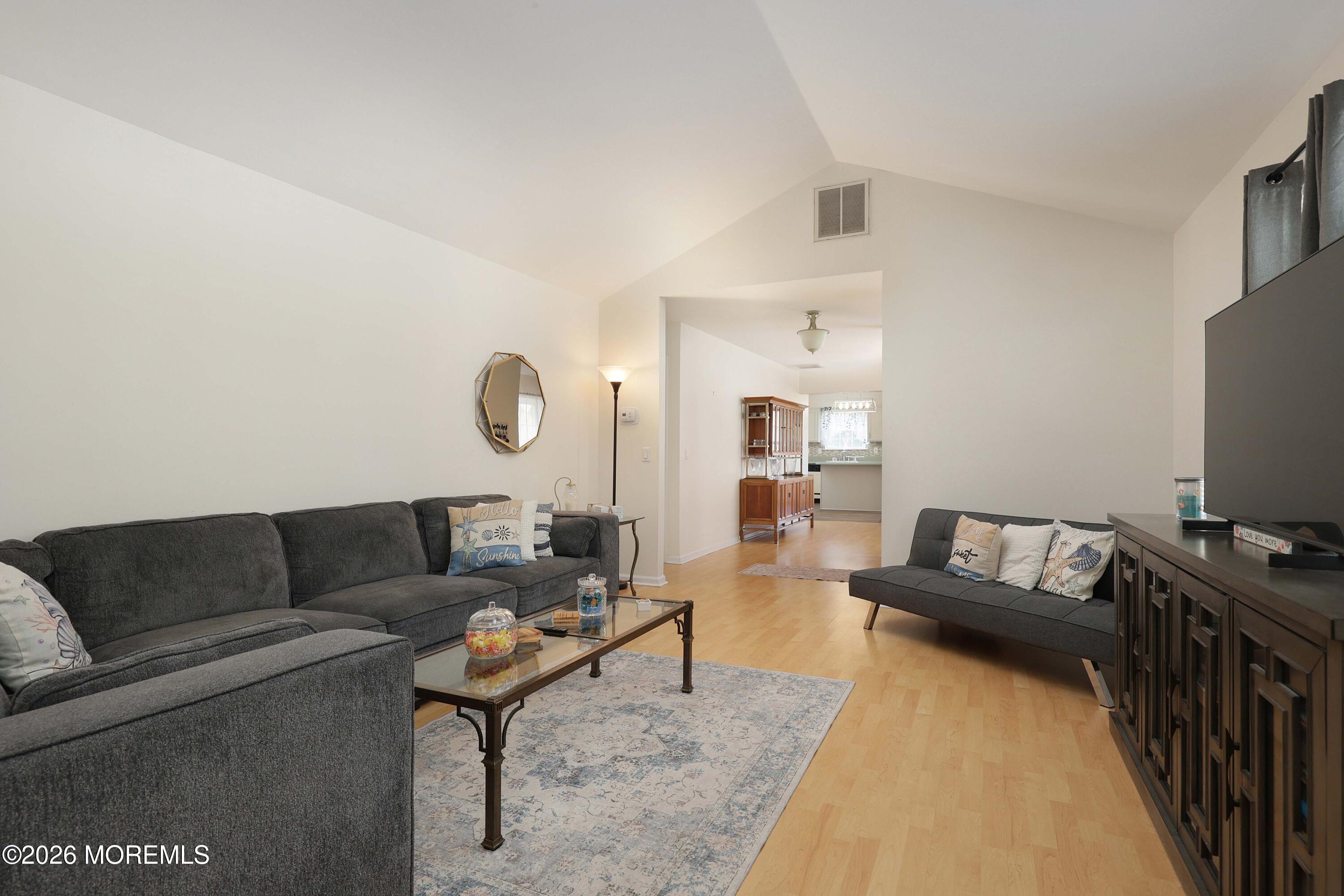 Living room with vaulted ceiling, gray sofas, glass coffee table, and doorway to the kitchen.