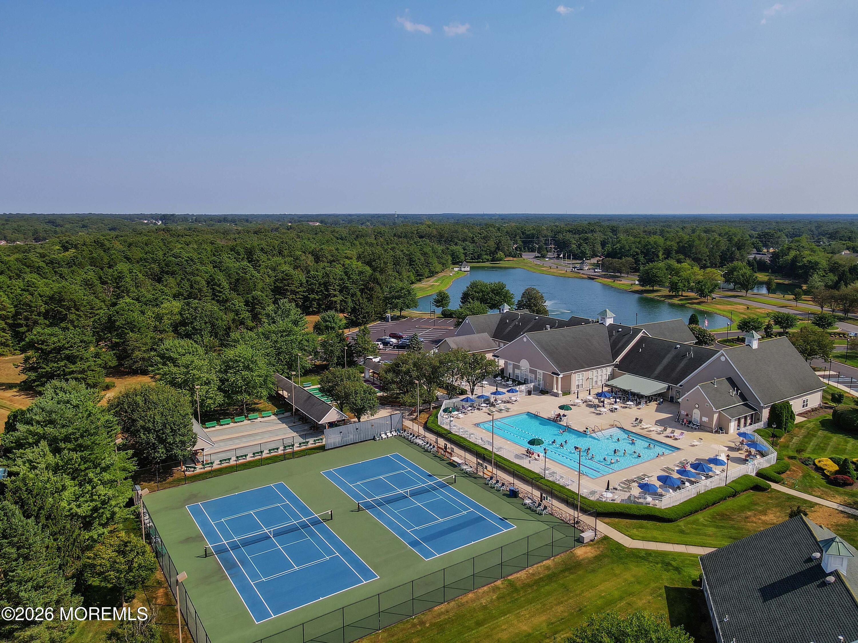 Aerial view of a community pool, tennis courts, clubhouse, and lake surrounded by trees.