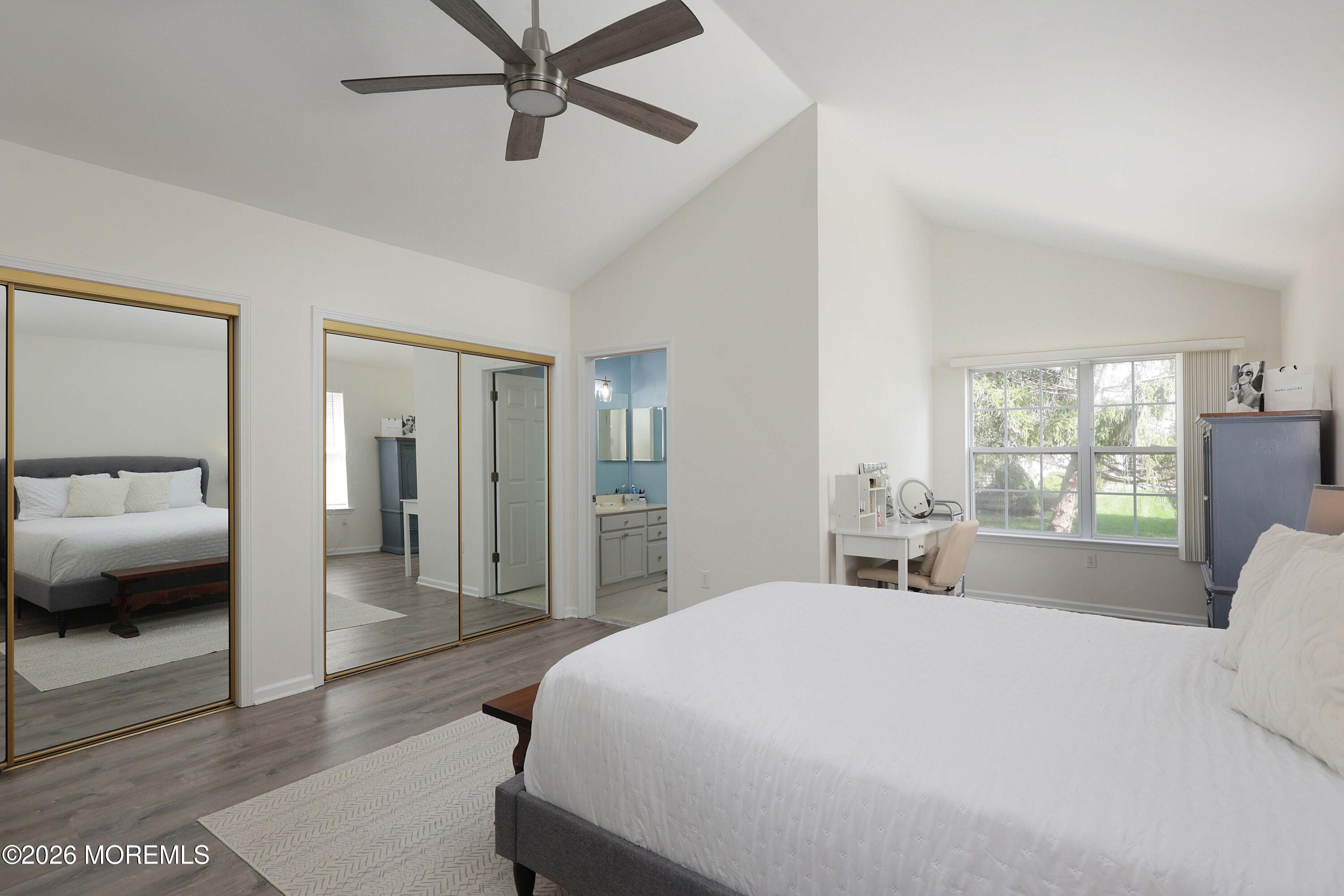 Primary bedroom with vaulted ceiling, mirrored closet doors, adjoining bathroom, and a desk beside a large window.