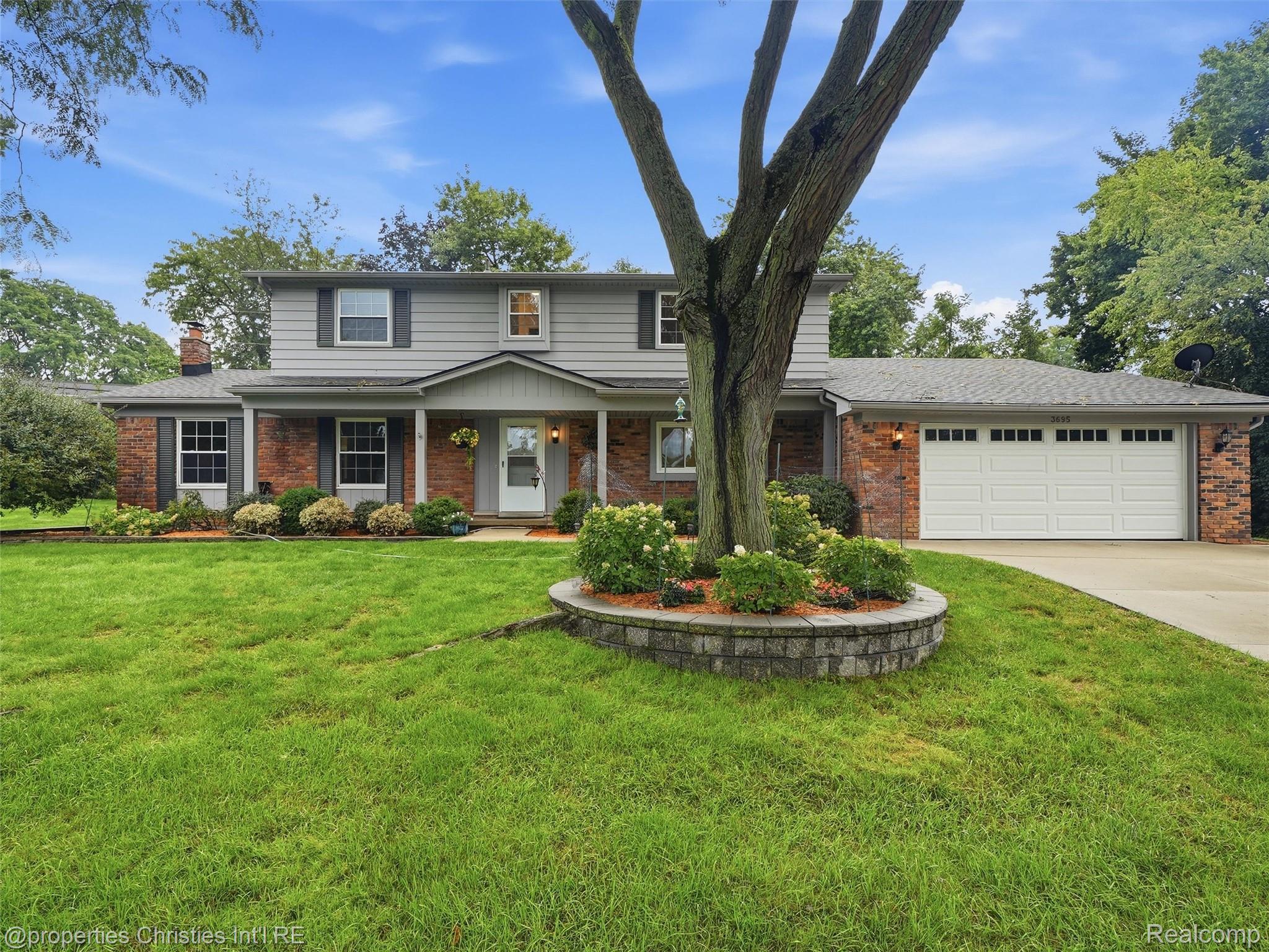 Two-story brick-and-siding home with a white garage, broad lawn, and mature tree in a raised planter.
