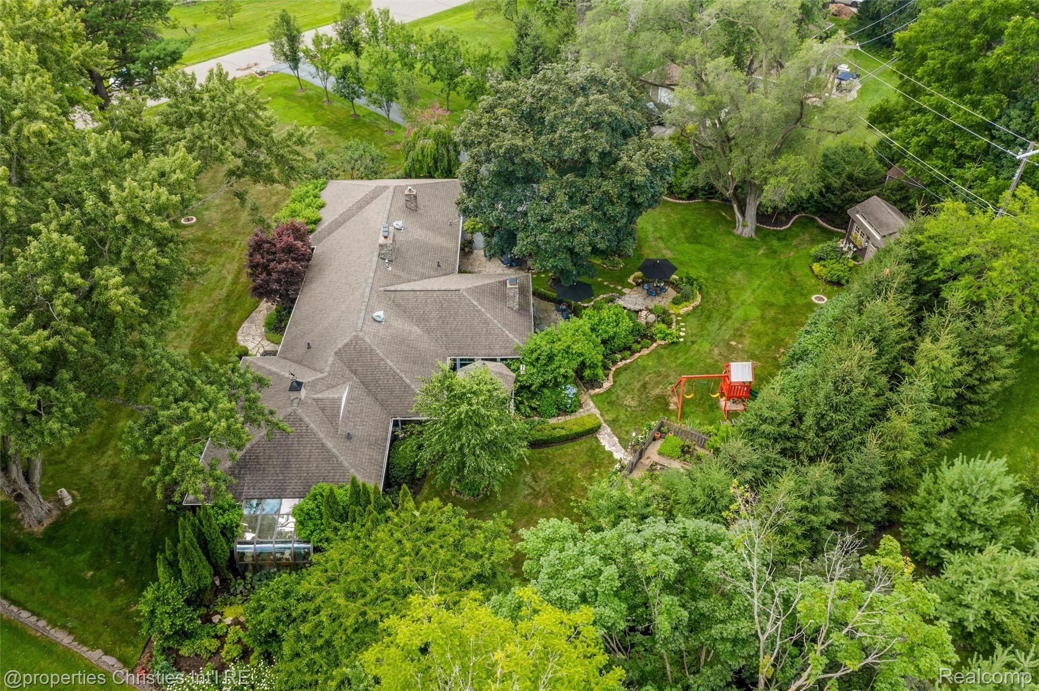 Aerial view of a tree-surrounded home with a landscaped yard, stone patio, and children’s playset.