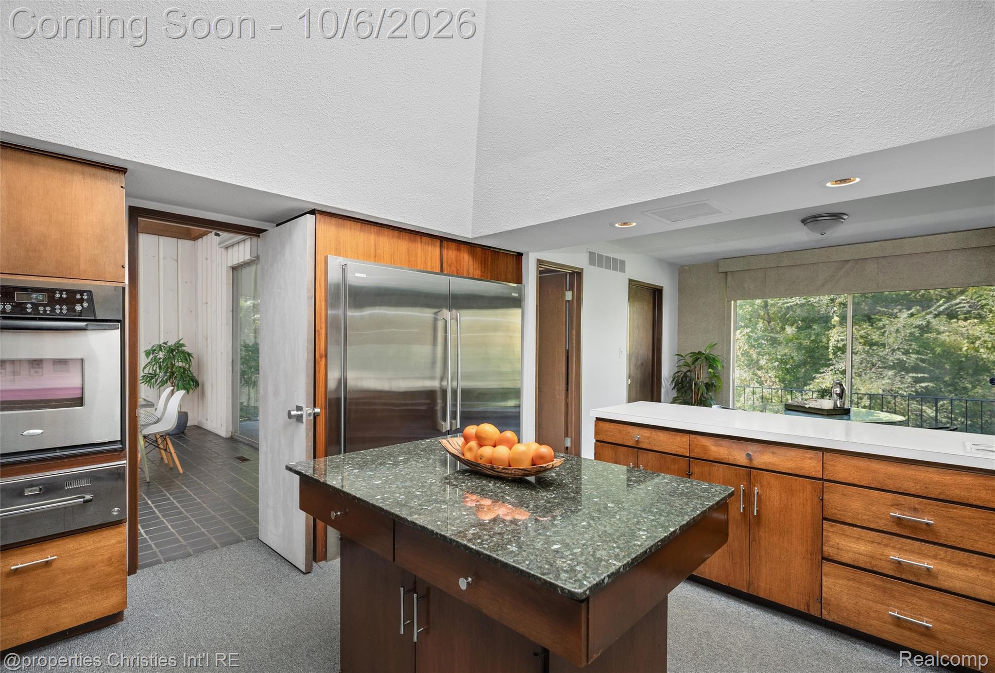 Kitchen with warm wood cabinetry, a dark stone island, stainless steel appliances, and a window overlooking trees.