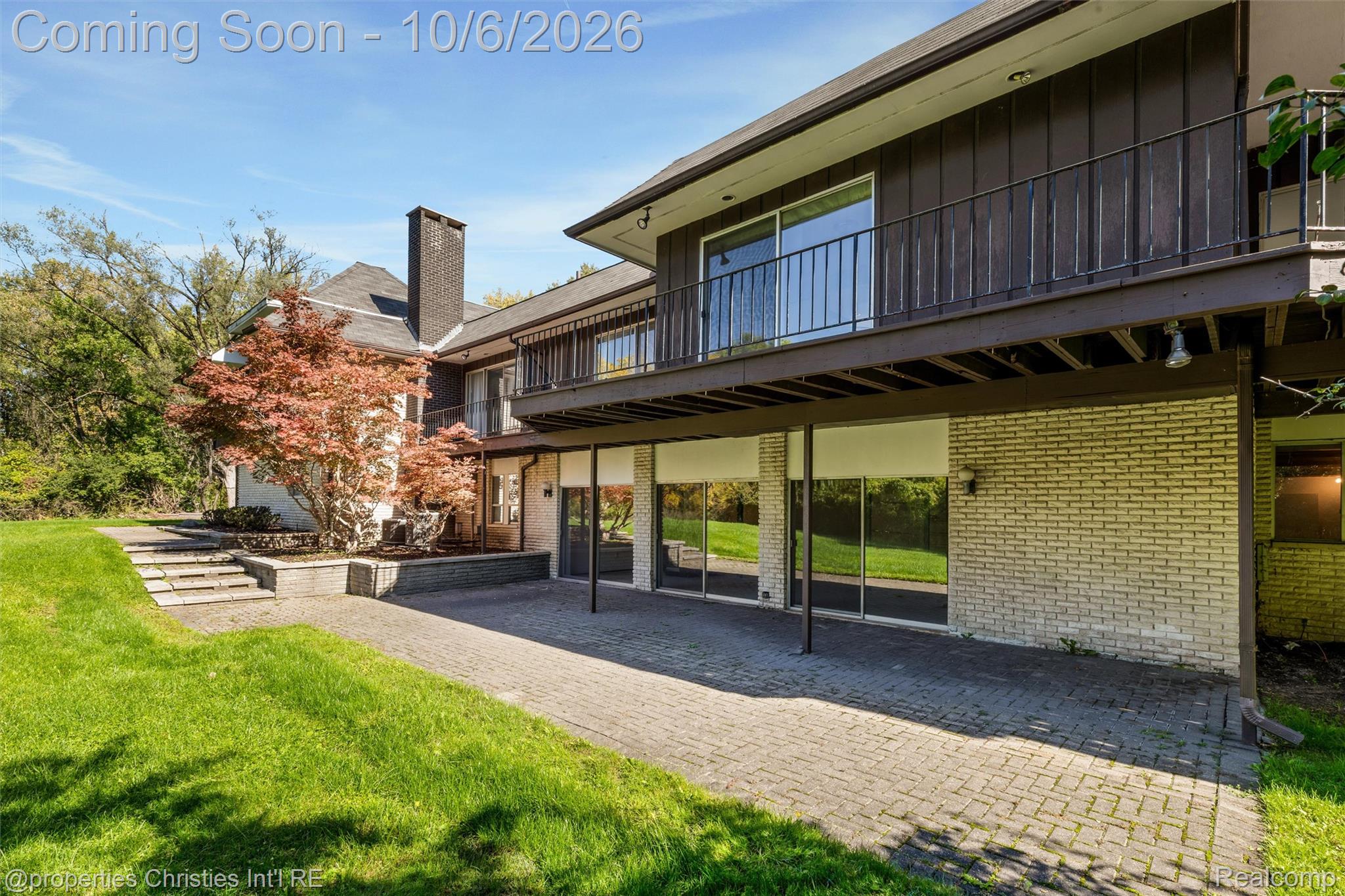 Home’s rear exterior with a raised deck overlooking a brick-paver patio and lawn.