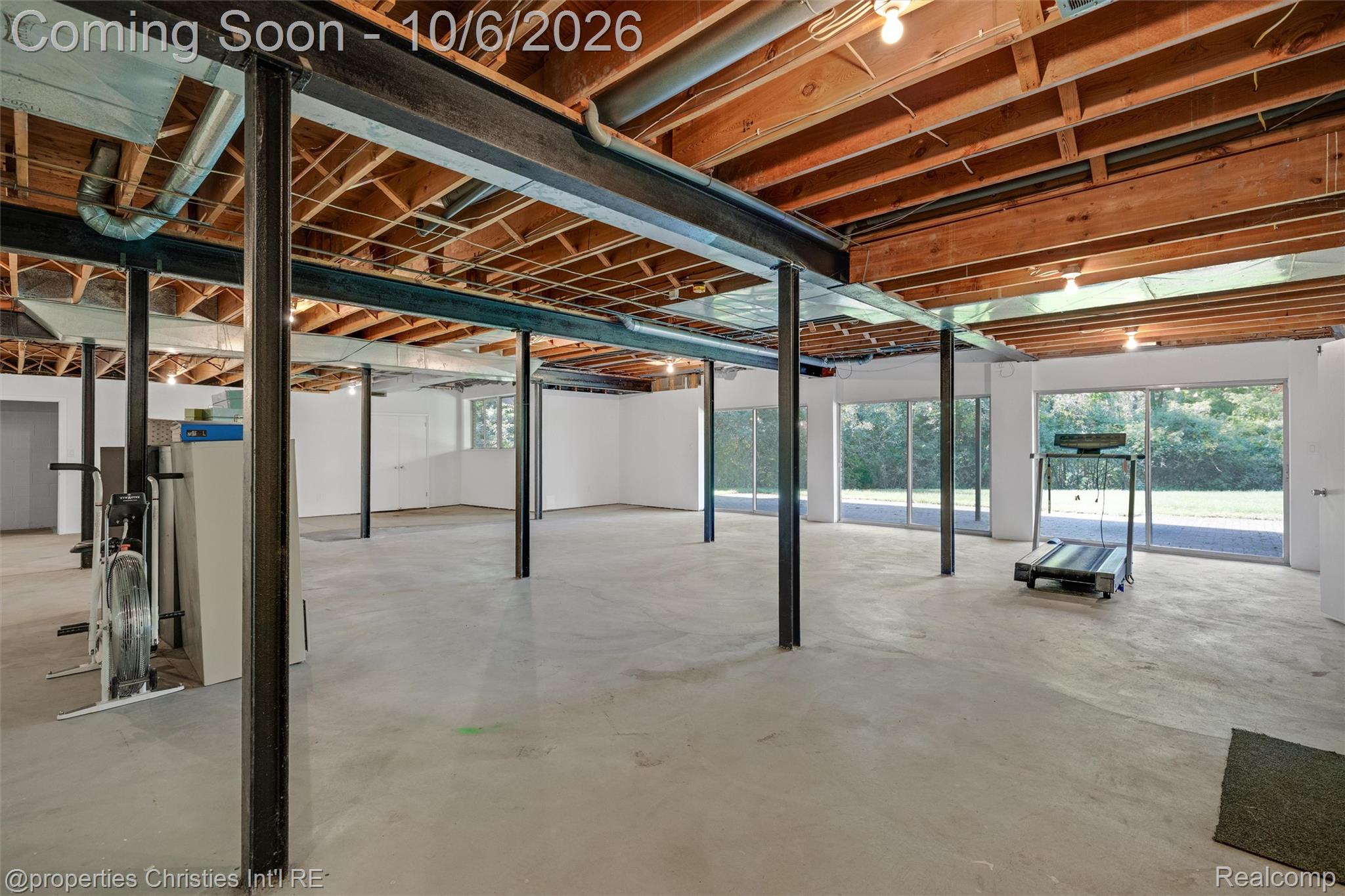 Open basement with exposed ceiling joists, steel support posts, and exercise equipment beside large windows.