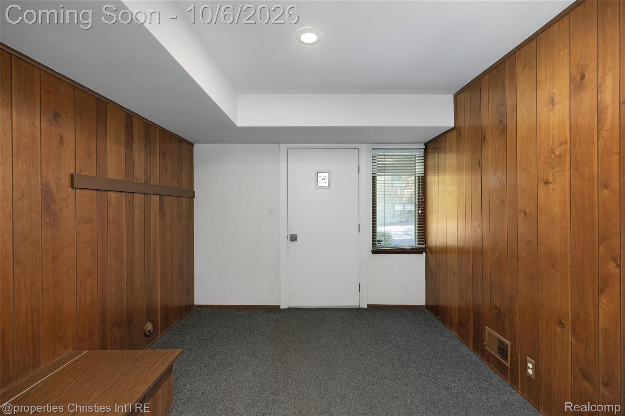 Entry room with wood-paneled walls, gray carpet, a white door, and a narrow window.