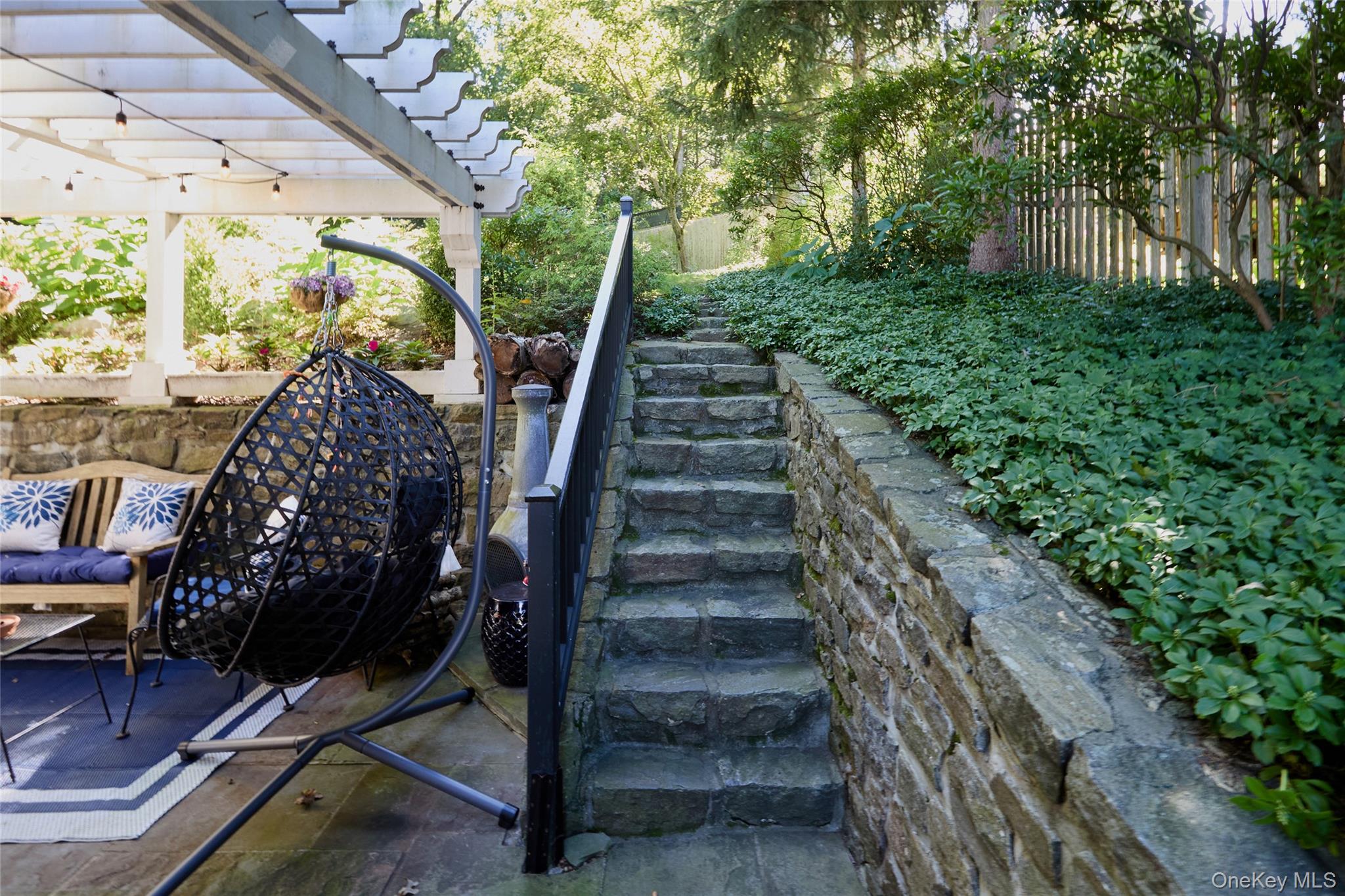 Furnished pergola patio with a hanging egg chair beside stone steps through a leafy garden.