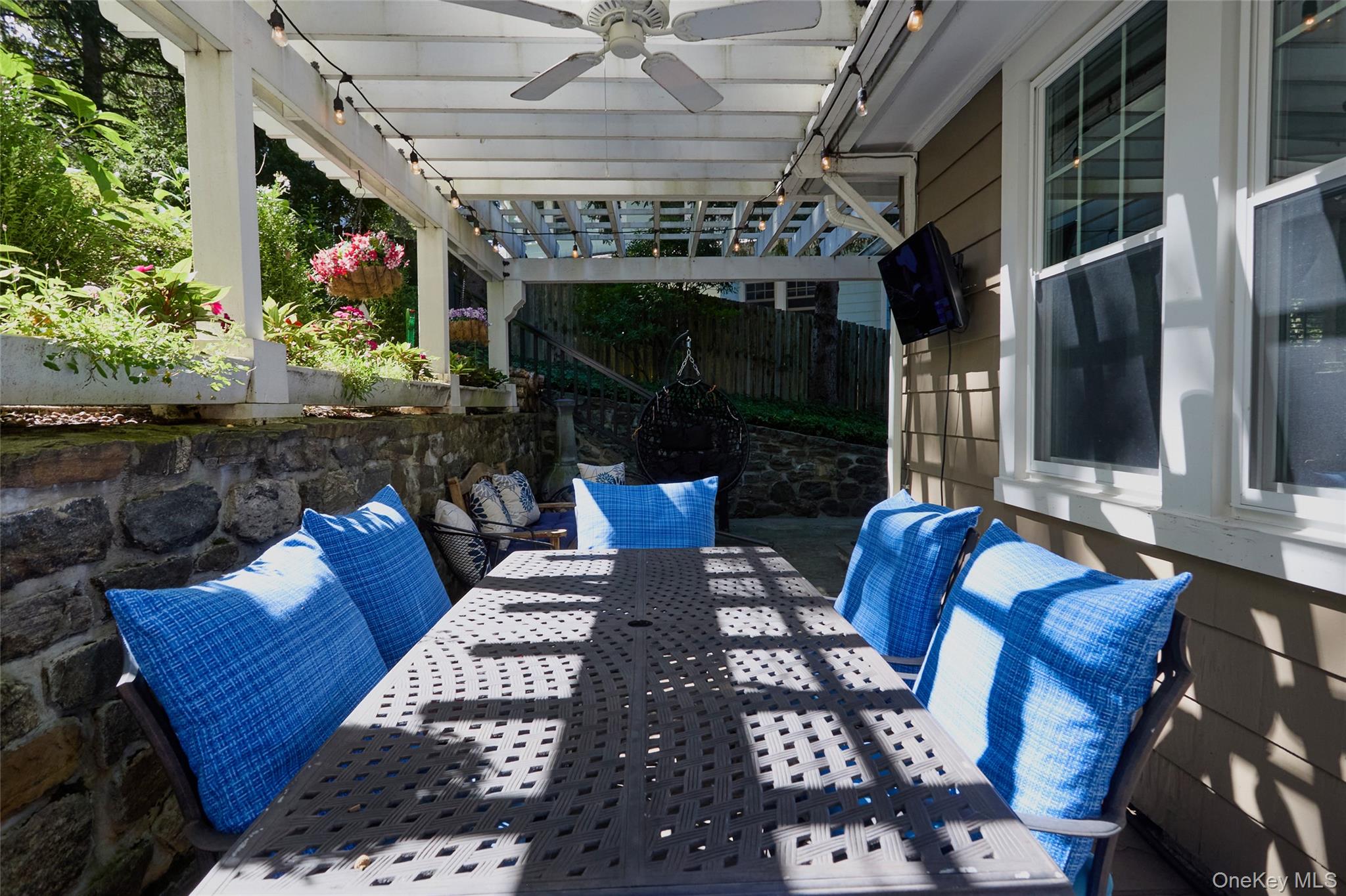 Covered patio with blue-cushioned dining chairs, a pergola, ceiling fan, and stone garden wall.