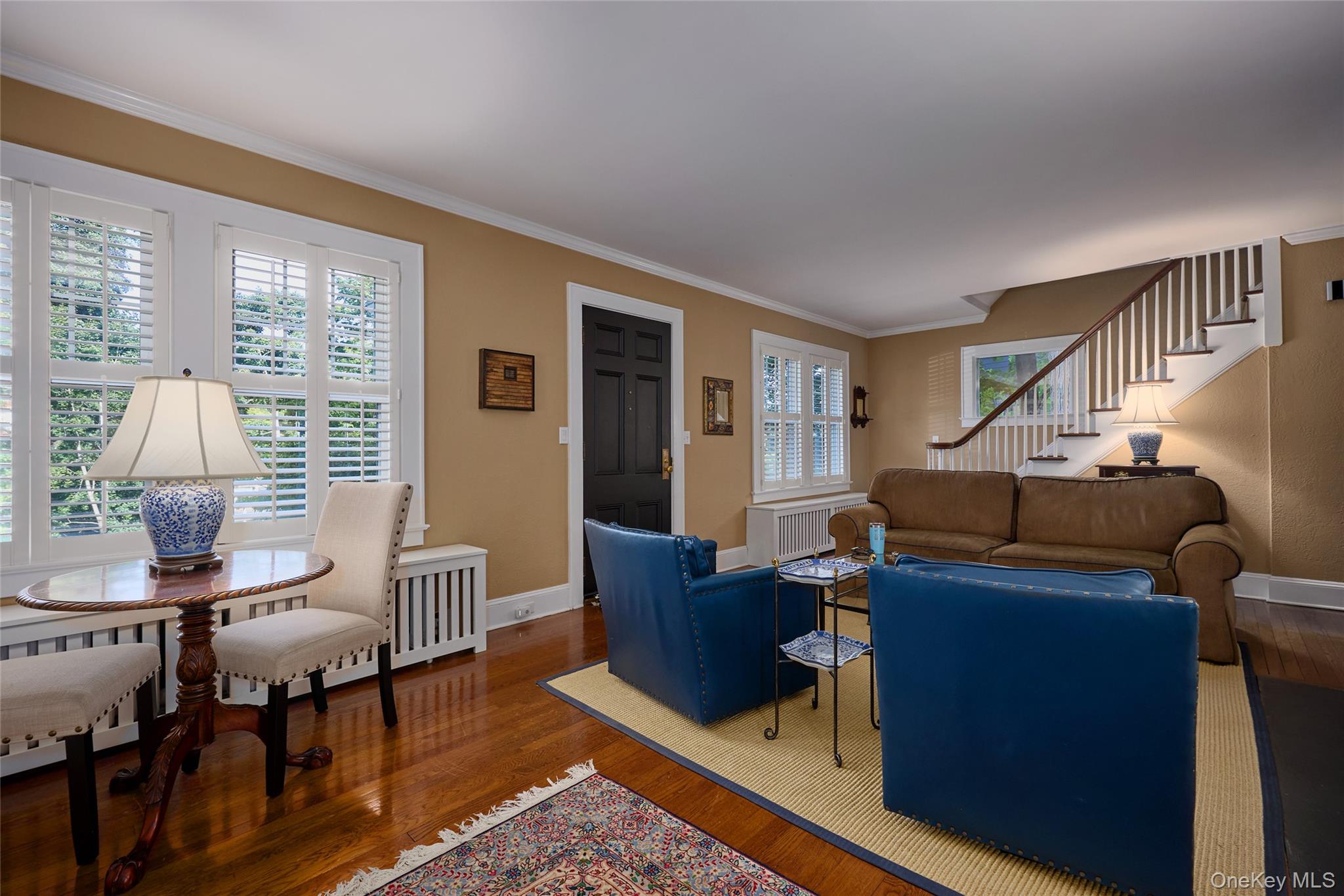 Living room with plantation shutters, hardwood floors, blue armchairs, and a staircase.