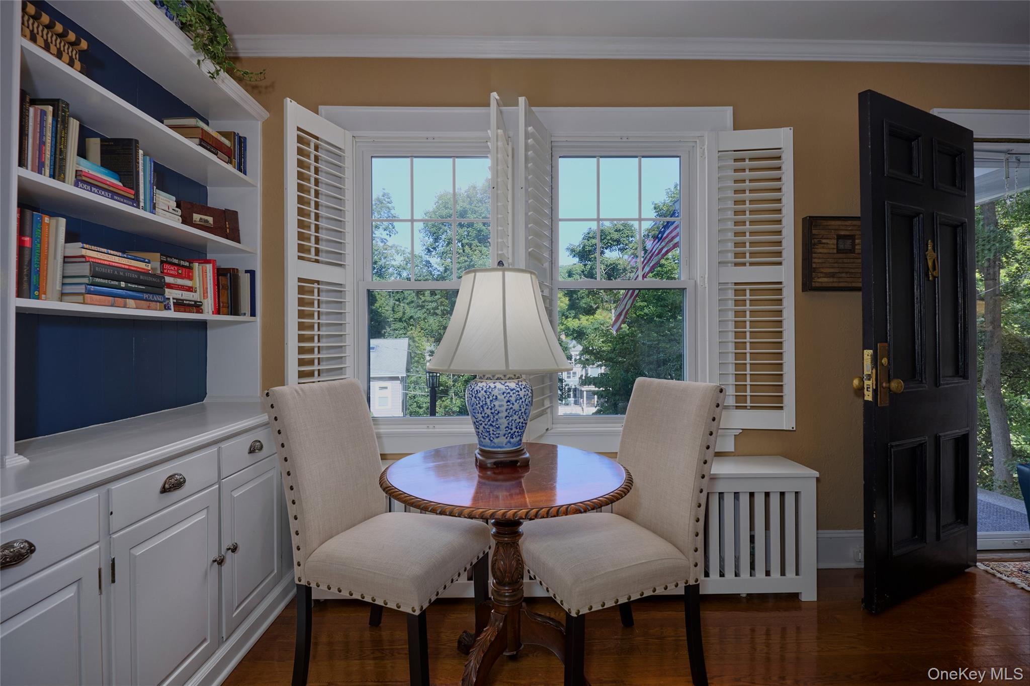 Dining nook with built-in bookshelves, a round table, two chairs, and shuttered windows overlooking trees.