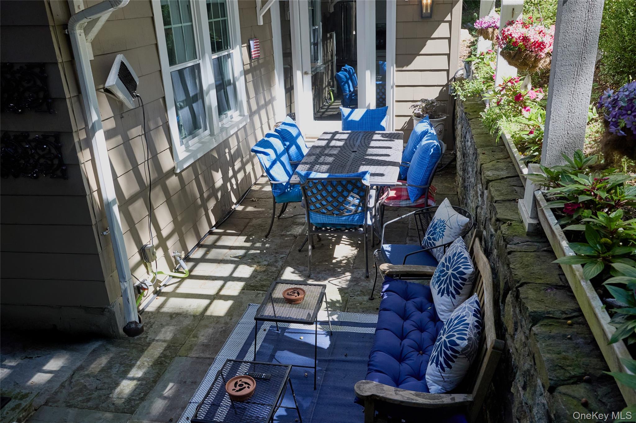 Furnished stone patio with blue outdoor seating, dining table, and colorful raised flower beds.