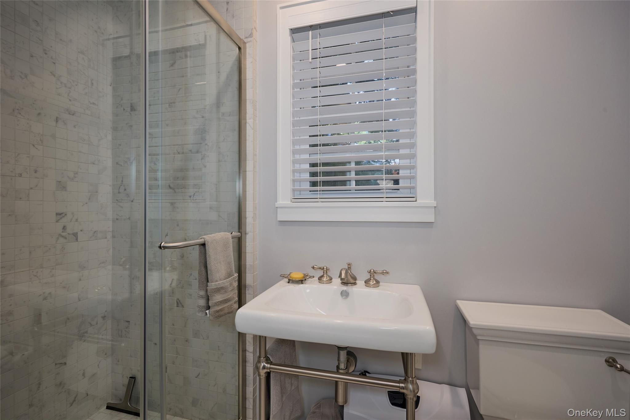 Bathroom with a marble-tiled glass shower, pedestal sink, and window with blinds.