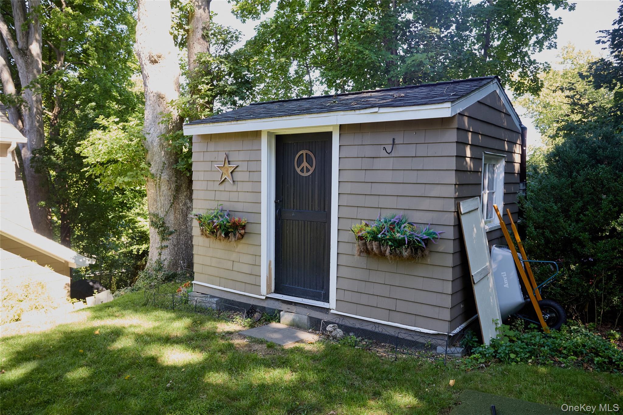 Gray-shingled shed with flower boxes and a peace-symbol door beneath mature trees.