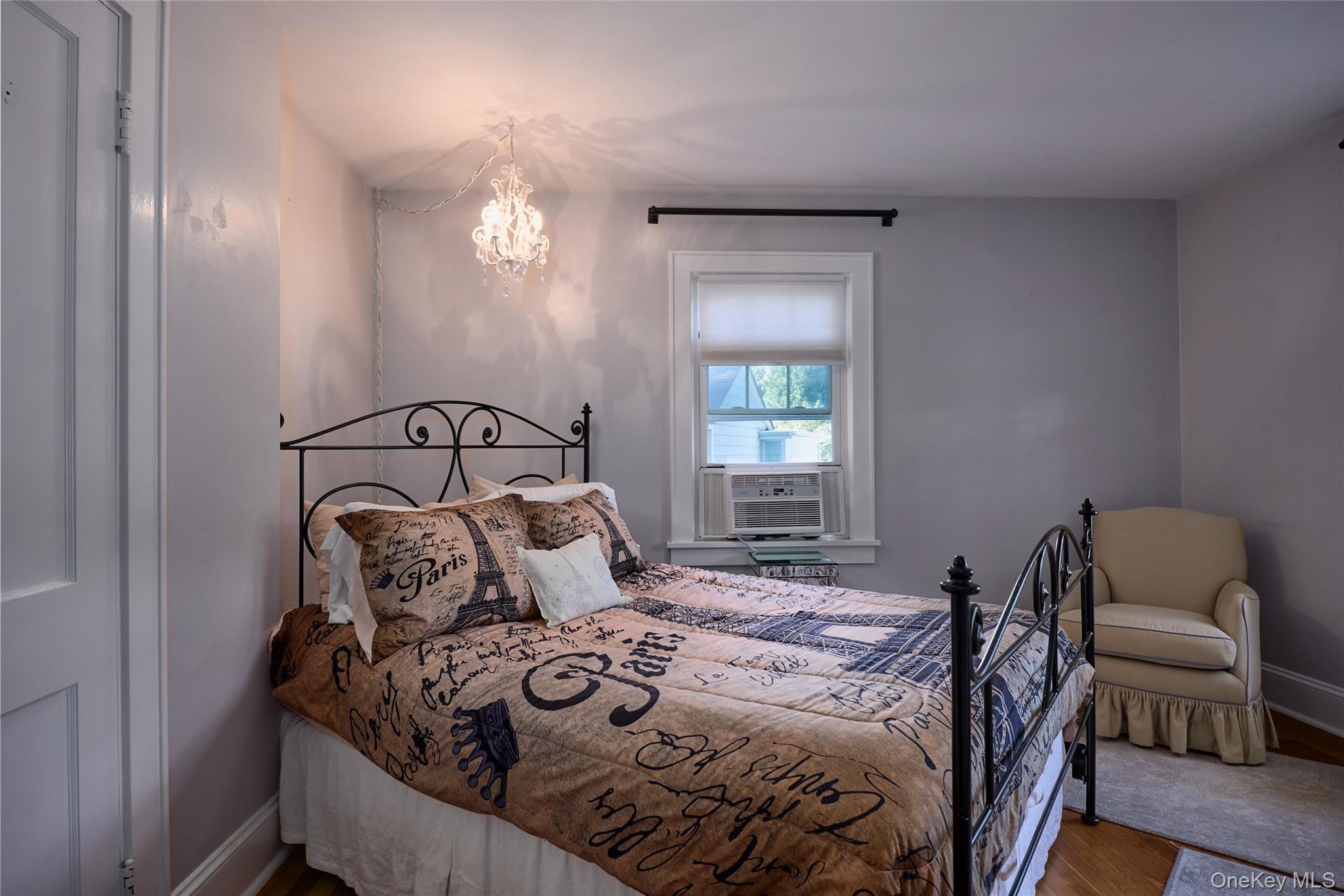 Bedroom with a black metal bed, Paris-themed bedding, crystal chandelier, and beige armchair.