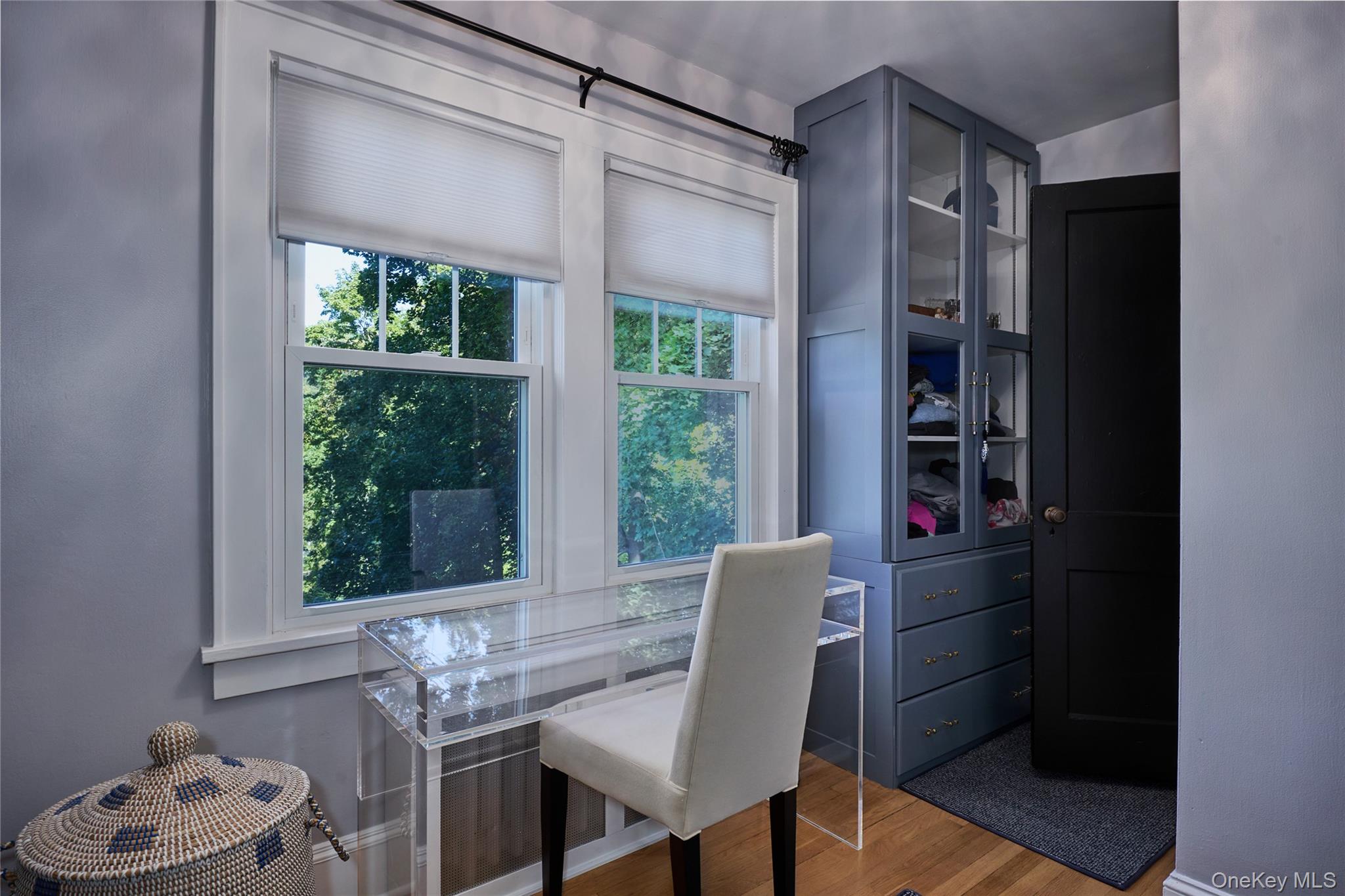 Furnished room with a clear desk, blue built-in cabinets, and windows overlooking trees.