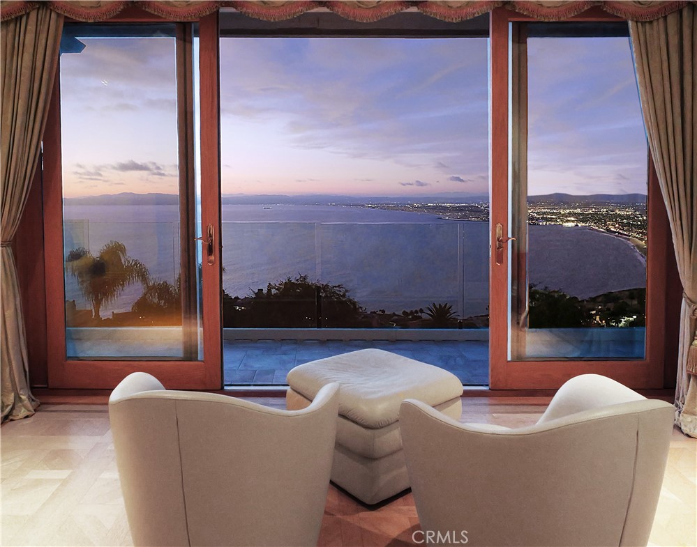 Living area with cream chairs facing glass doors and a balcony overlooking the ocean at dusk.