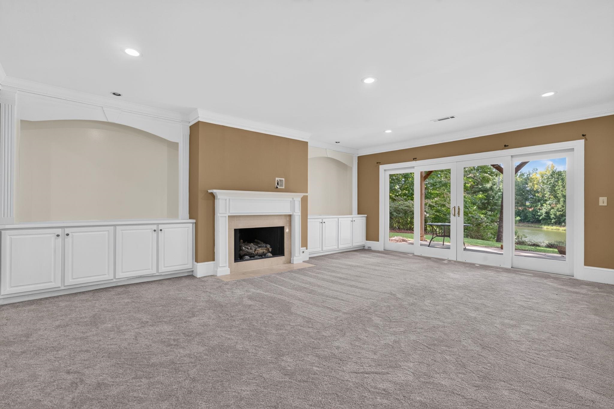 Empty living room with a white fireplace, built-in cabinets, and glass doors overlooking a pond.