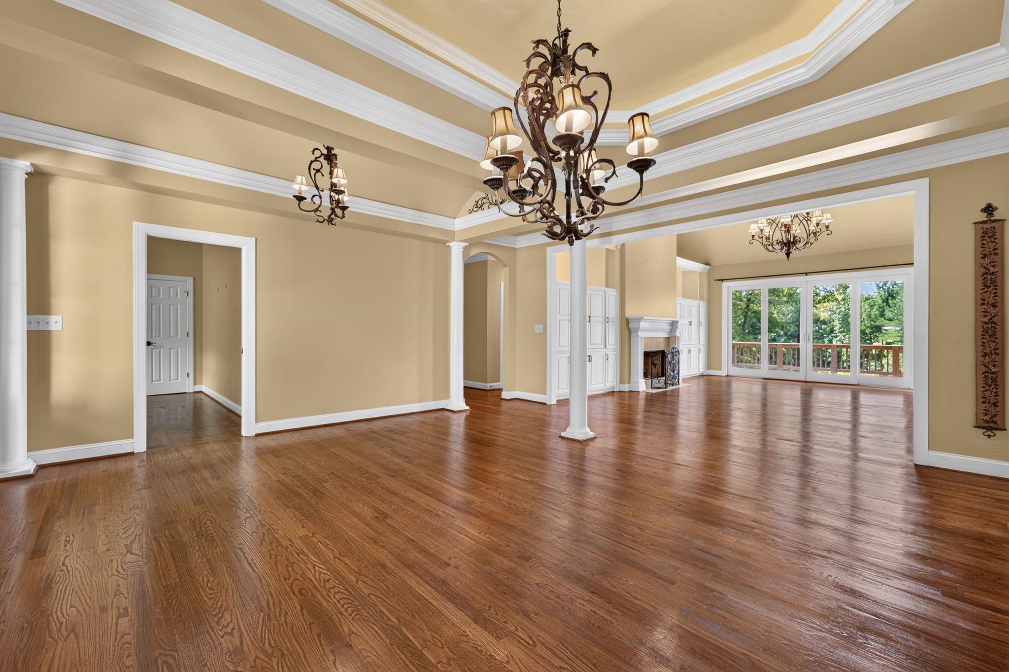 Open living and dining area with hardwood floors, ornate chandeliers, and glass doors to a leafy deck.
