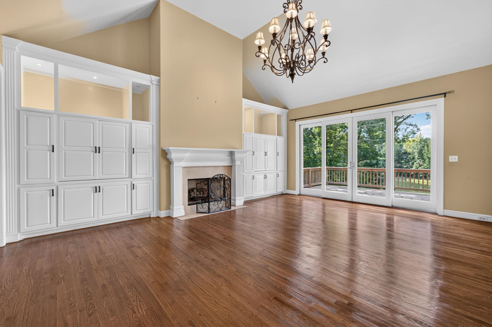 Living room with vaulted ceiling, hardwood floors, white built-ins, fireplace, and glass doors to a tree-lined deck.
