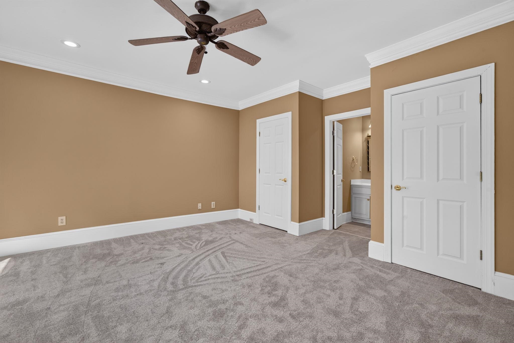 Empty bedroom with tan walls, gray carpet, and an open doorway to the bathroom.