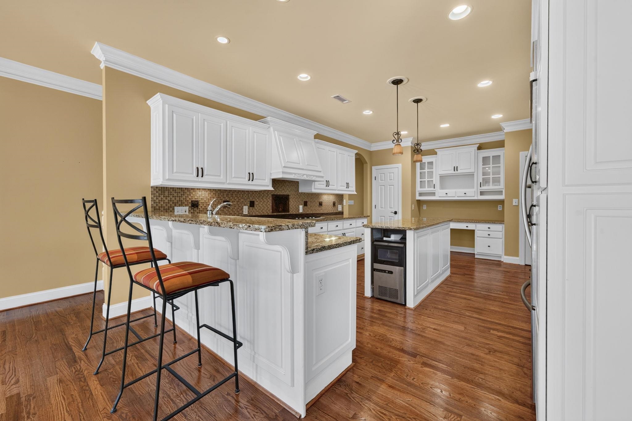 Kitchen with white cabinets, granite island, two bar stools, and warm hardwood floors.