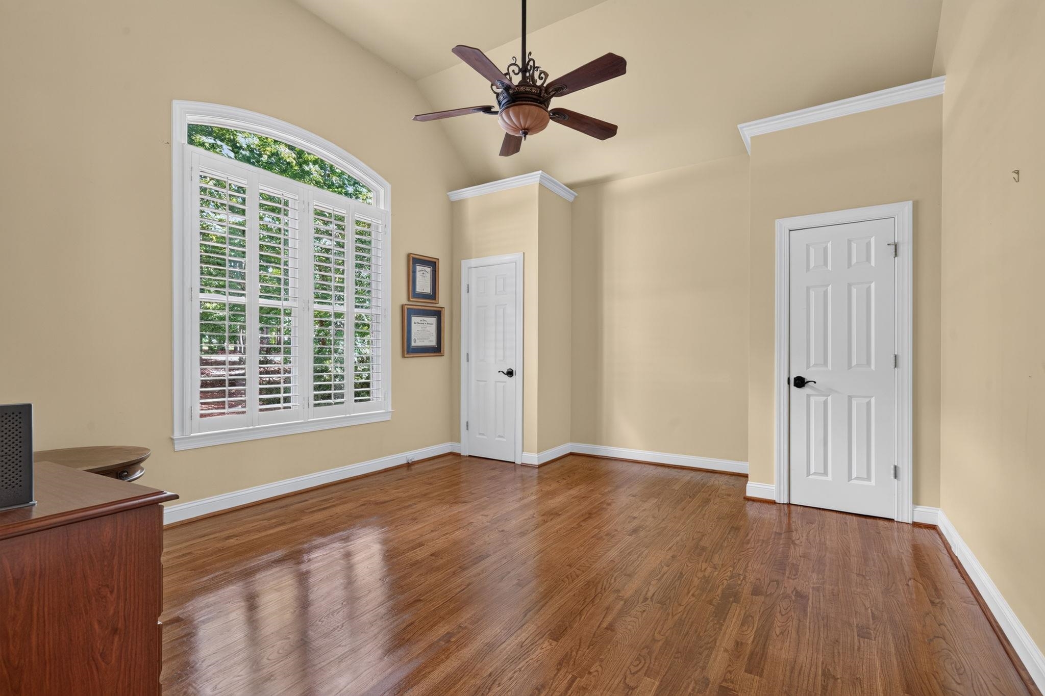 Room with hardwood floors, an arched shuttered window, and a high ceiling with a ceiling fan.