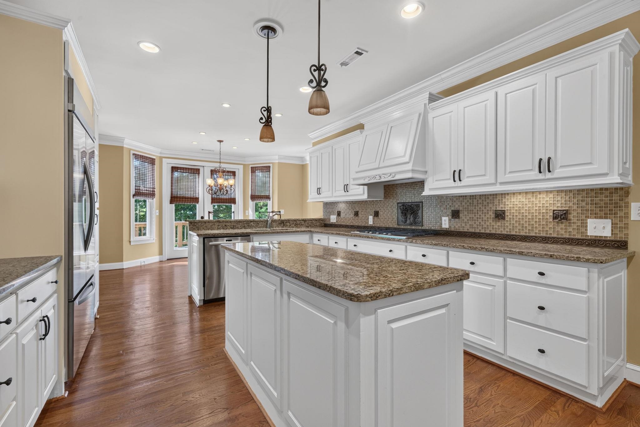 Kitchen with white cabinets, granite island, mosaic backsplash, and tall windows with woven shades.