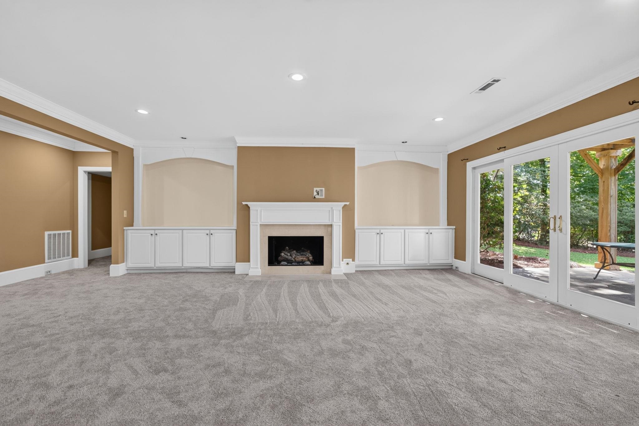 Empty living room with a fireplace, white built-in cabinets, gray carpet, and glass doors to a patio.