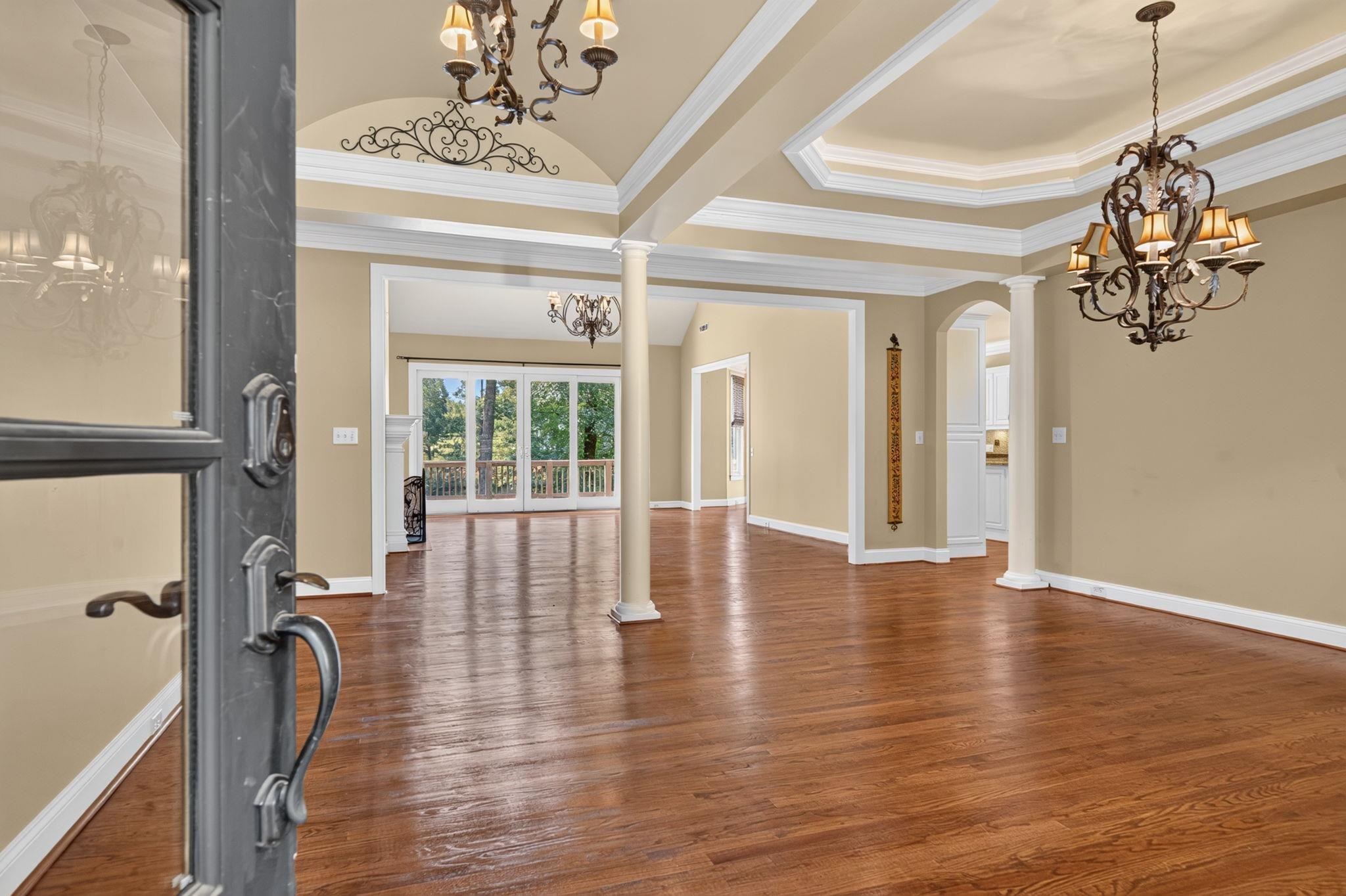 Vacant formal room with hardwood floors, ornate chandeliers, columns, and glass doors to a deck.