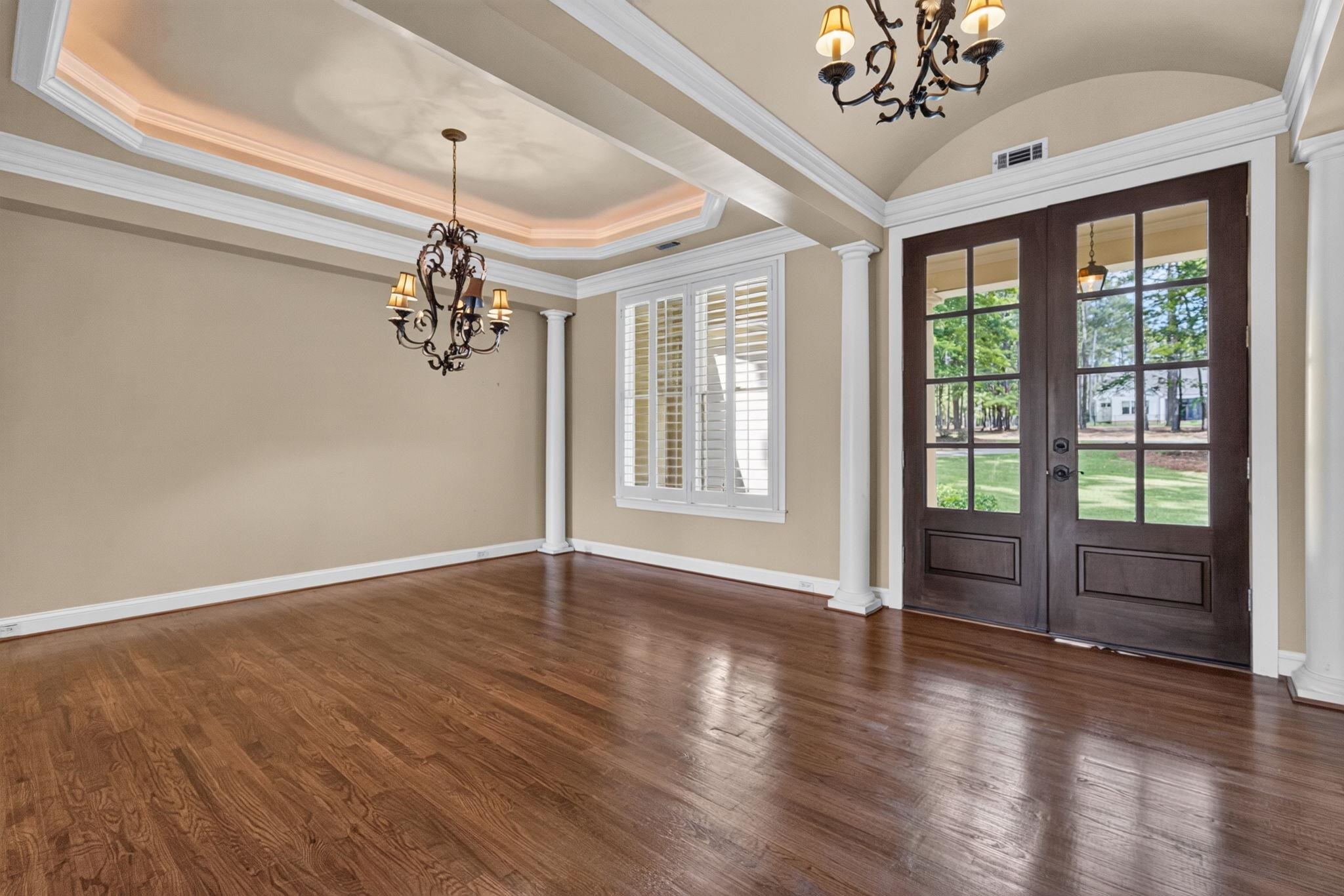 Empty dining room with hardwood floors, tray ceiling, chandeliers, and French doors to the lawn.