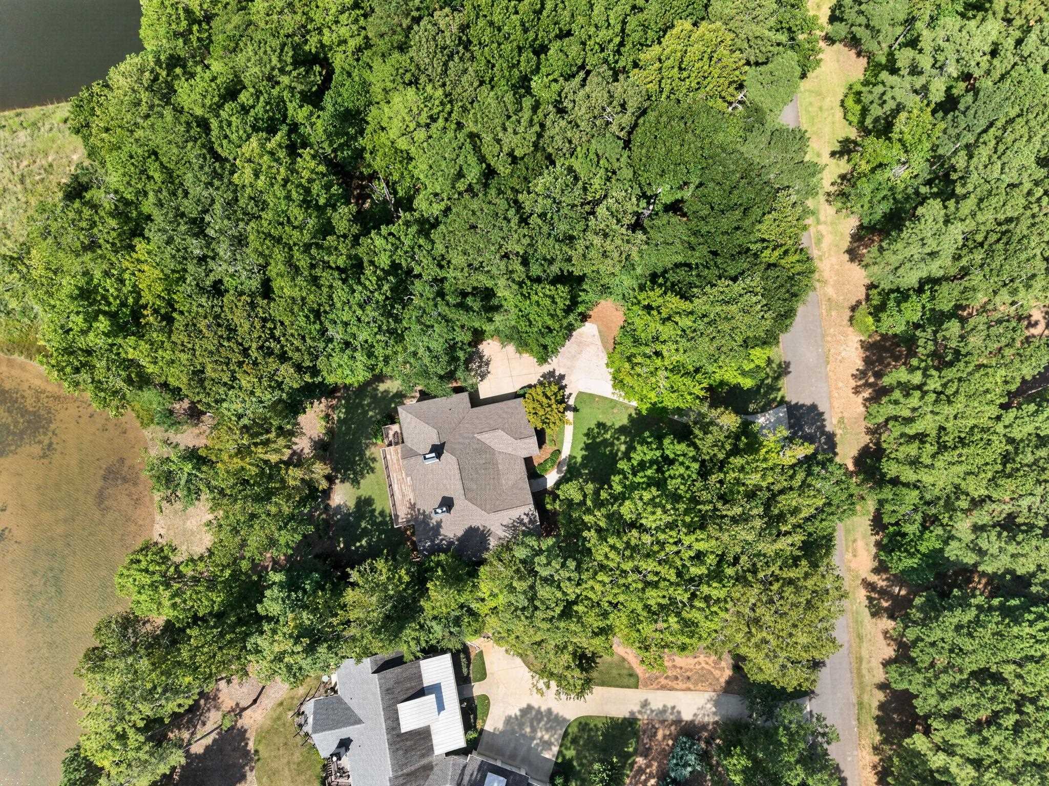 Aerial view of a wooded home beside a pond, with a curving driveway and neighboring house.