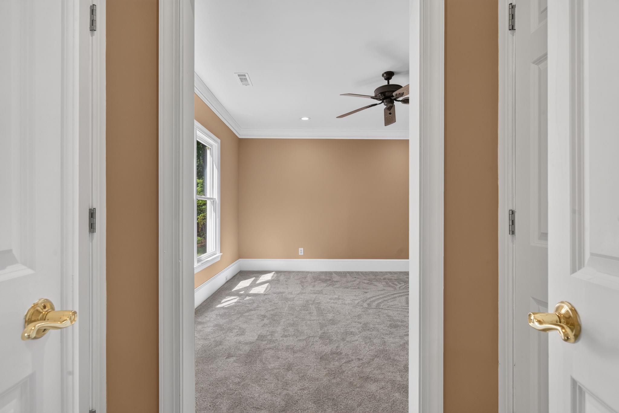Empty bedroom with gray carpet, tan walls, a ceiling fan, and a sunlit window.