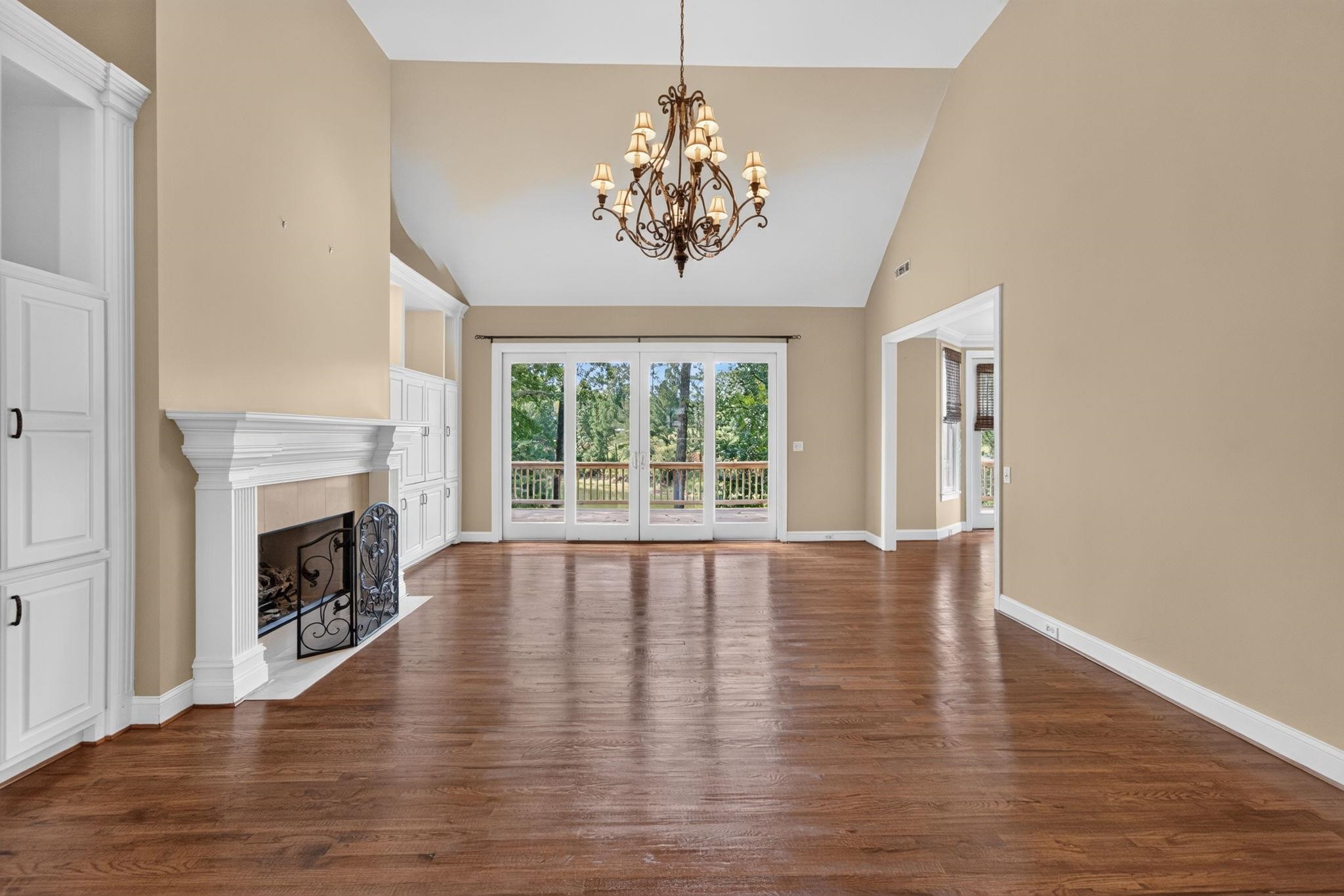 Empty living room with hardwood floors, a white fireplace, and glass doors opening to a tree-lined deck.
