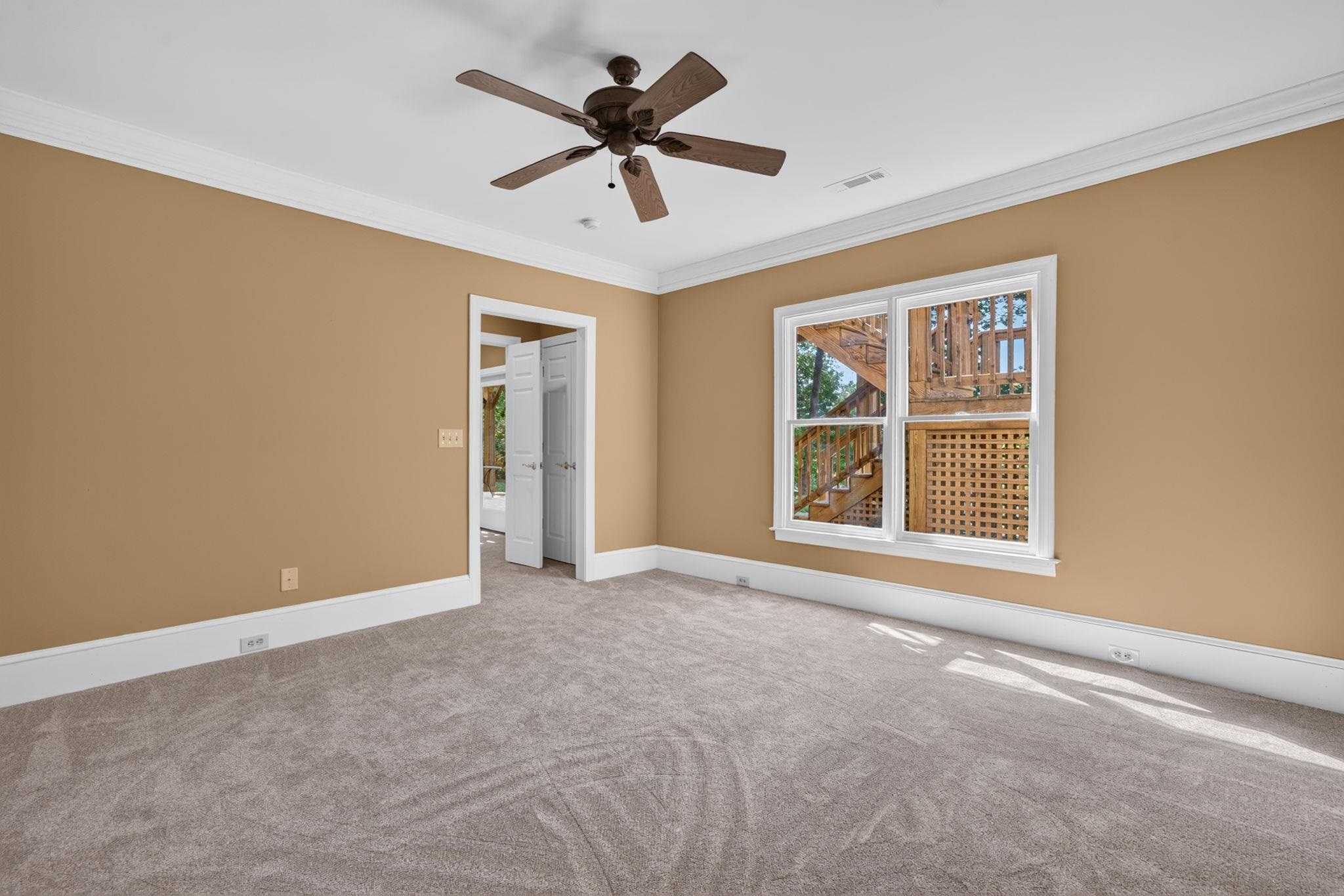 Empty bedroom with beige carpet, a ceiling fan, and a window overlooking a wooden deck.