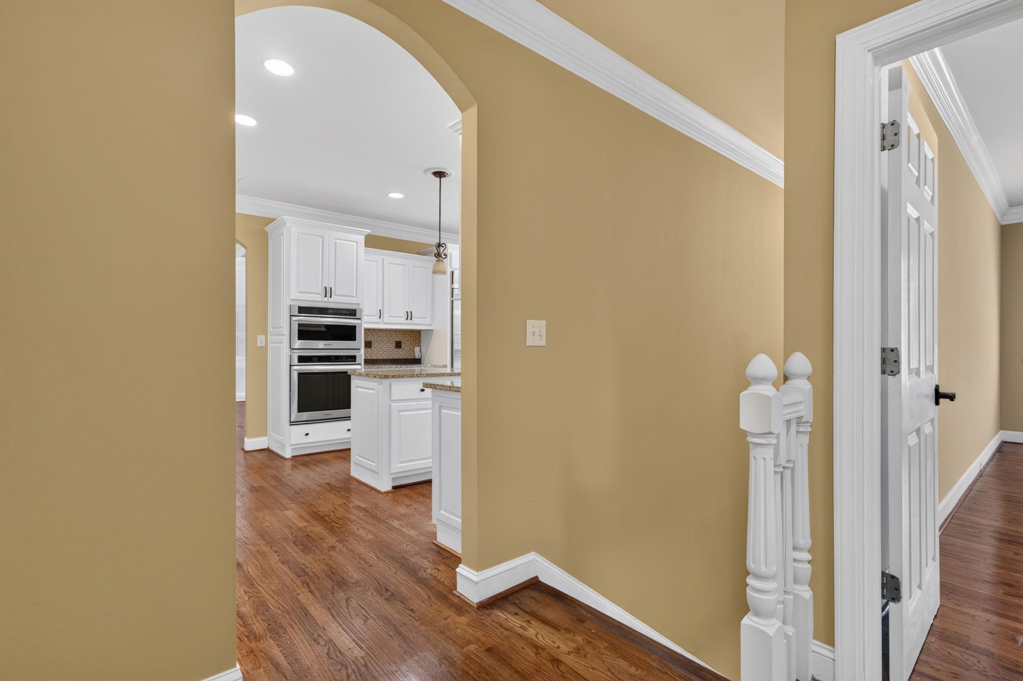 Hallway with hardwood floors and an arched opening into a white kitchen with double wall ovens.