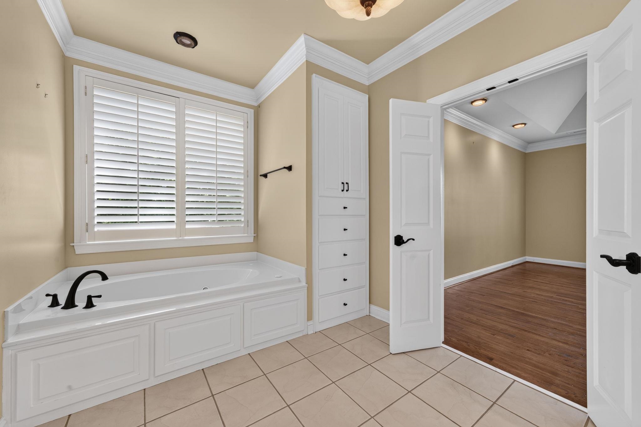 Bathroom with a built-in soaking tub beneath white plantation shutters and an open doorway to hardwood flooring.