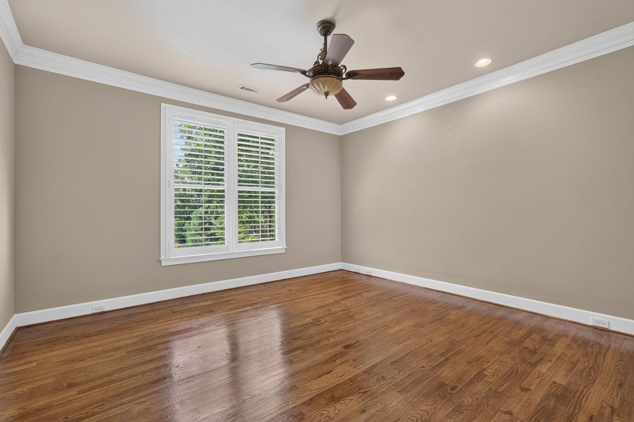 Empty bedroom with hardwood floors, a ceiling fan, and plantation shutters overlooking greenery.
