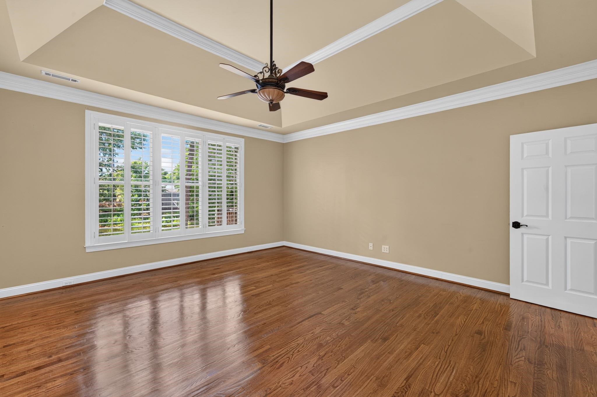 Empty room with polished hardwood floors, plantation shutters, and a tray ceiling with ceiling fan.