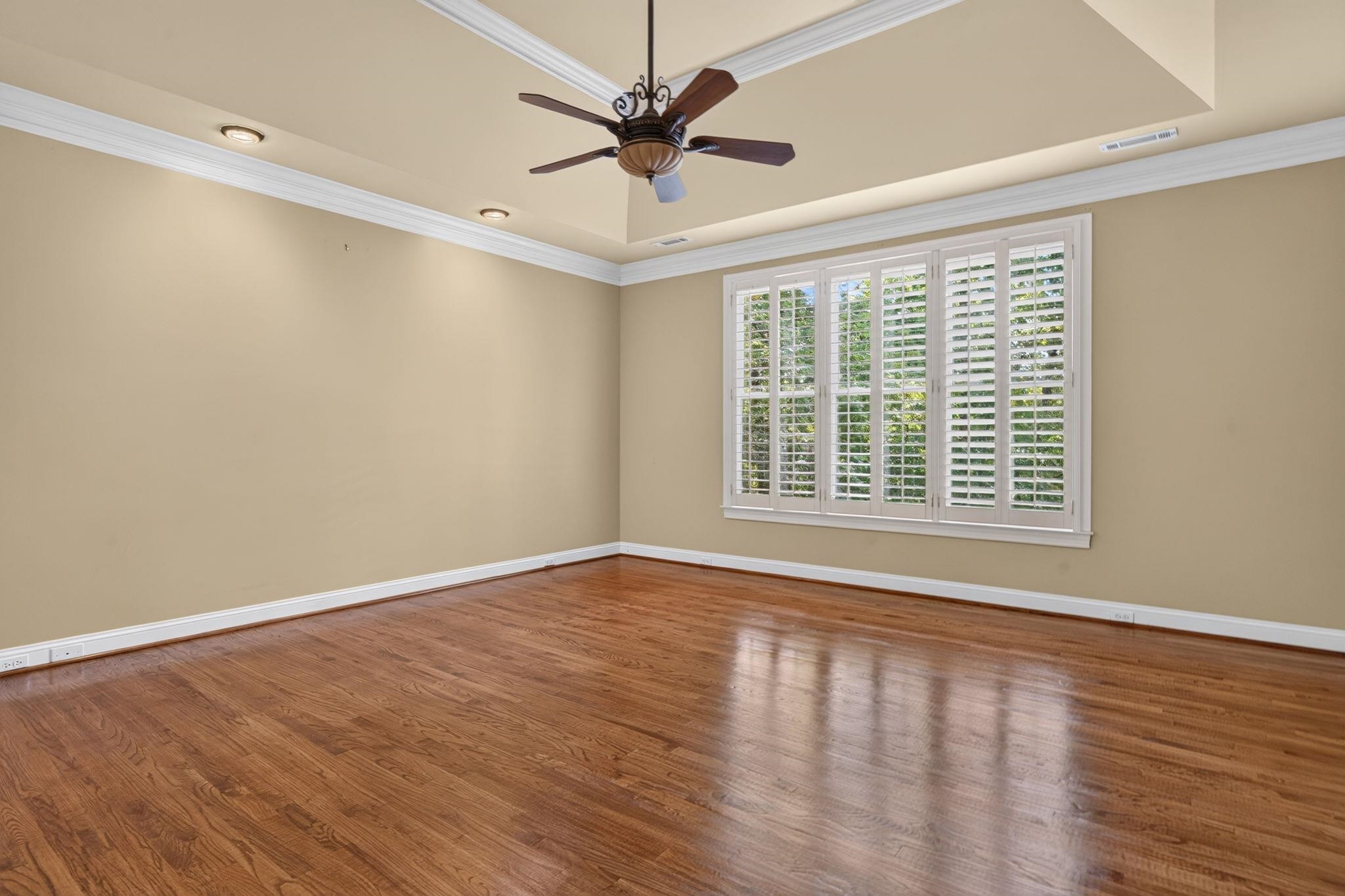 Empty bedroom with hardwood floors, a tray ceiling, and wide plantation-shuttered window.