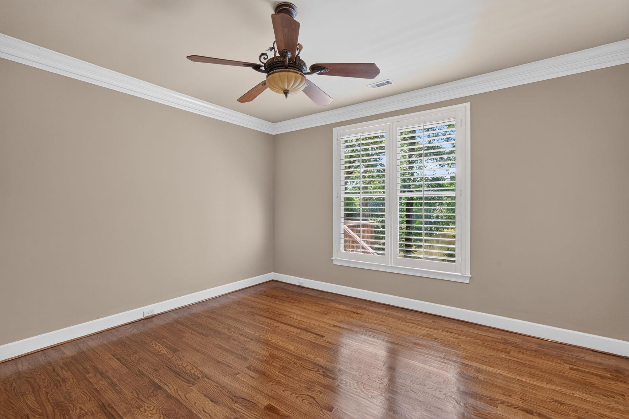 Empty bedroom with hardwood floors, plantation-shuttered window, and ceiling fan.