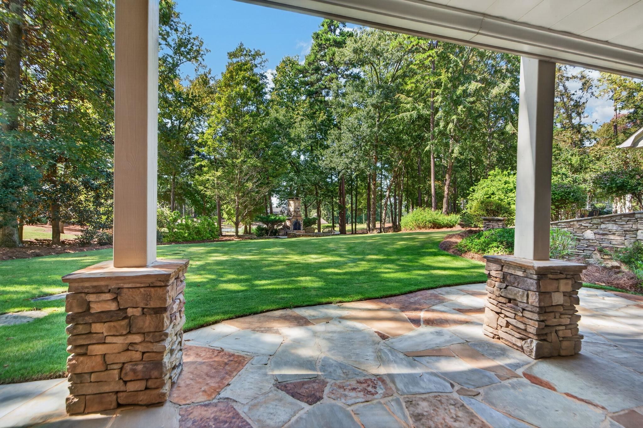 Covered stone patio overlooking a green lawn and wooded backyard, with an outdoor fireplace in the distance.