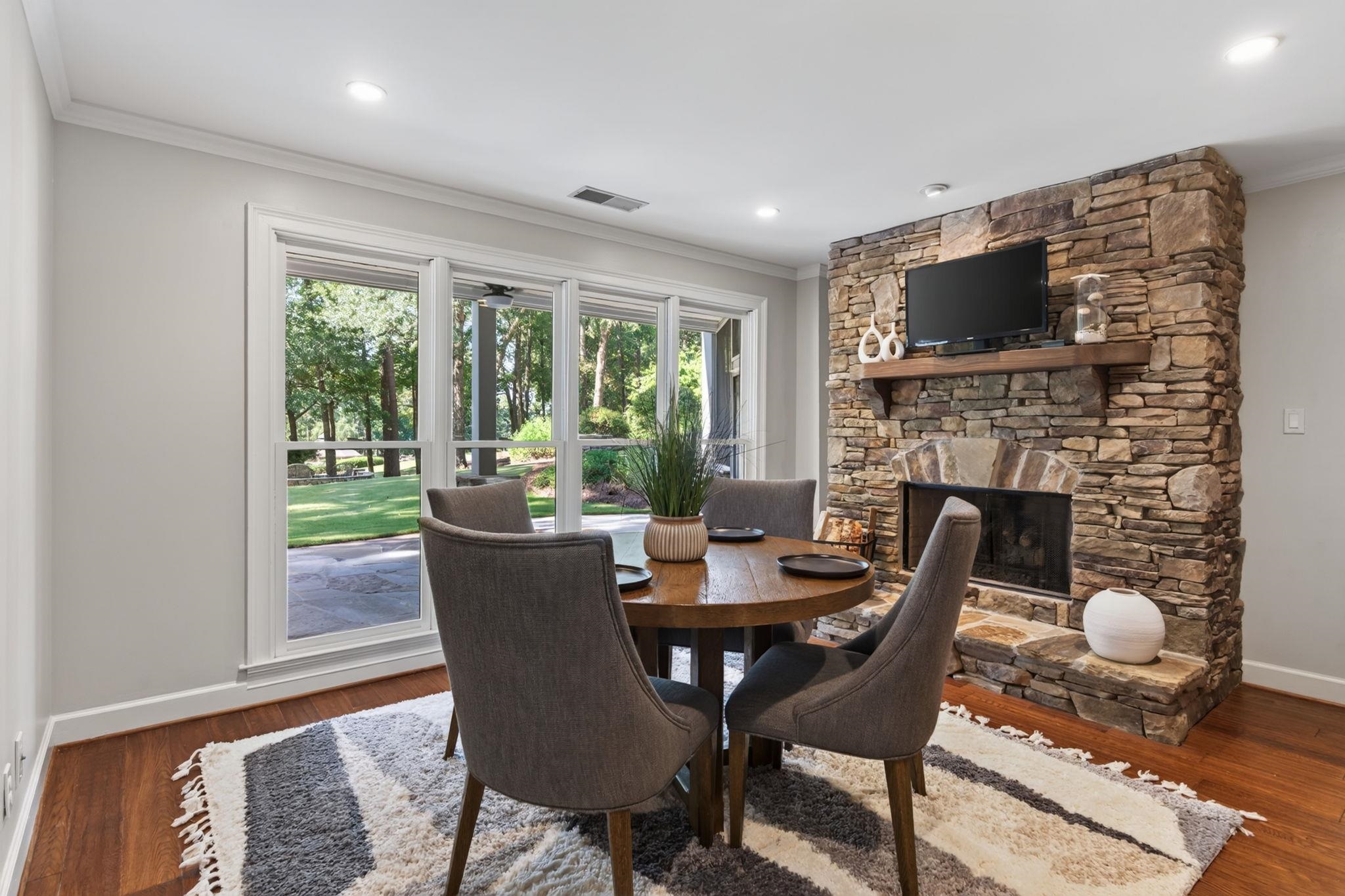 Dining area with a stone fireplace, round table, and large windows overlooking a leafy lawn.