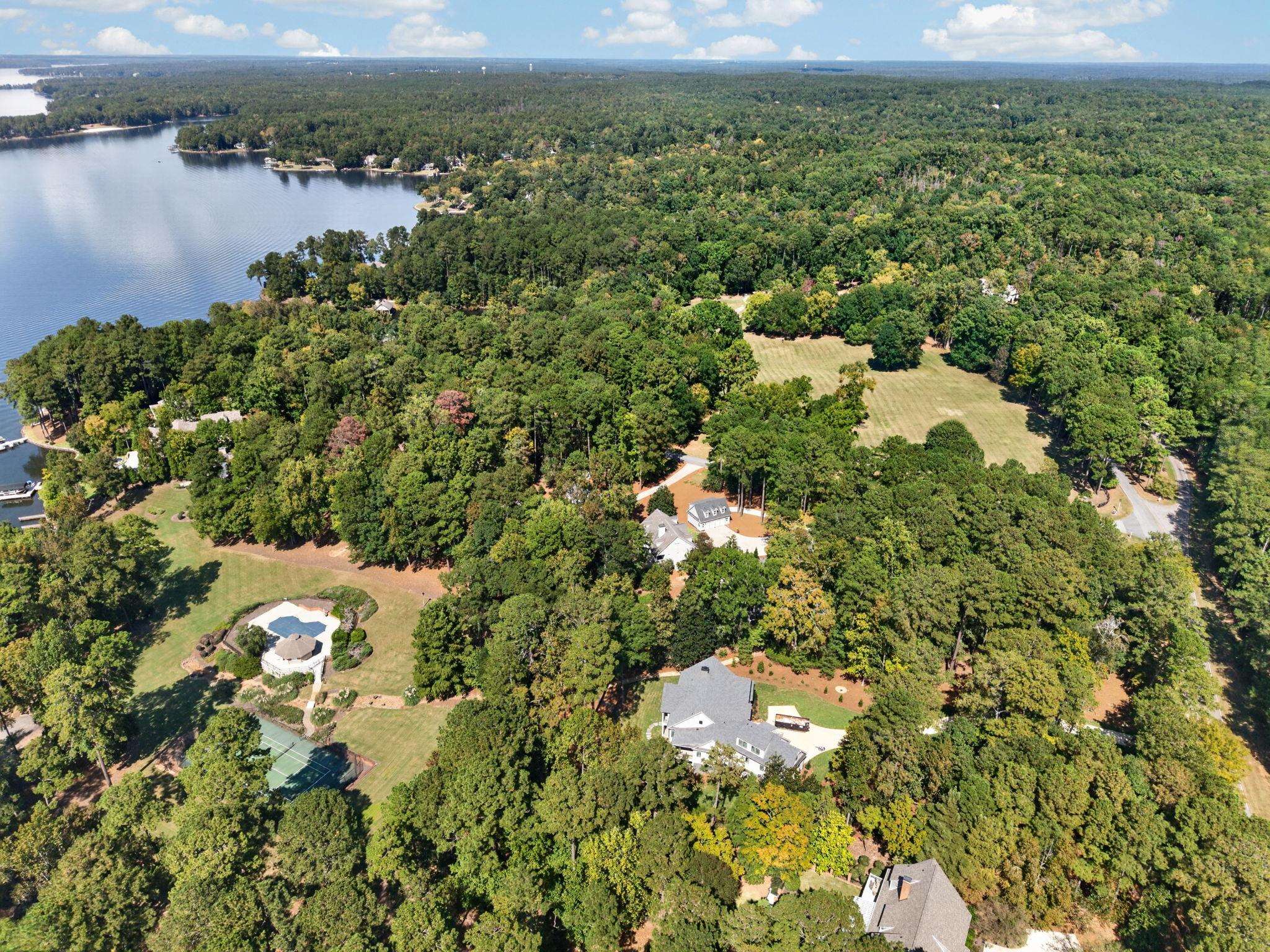 Aerial view of wooded homes beside a lake, with a pool and tennis court.