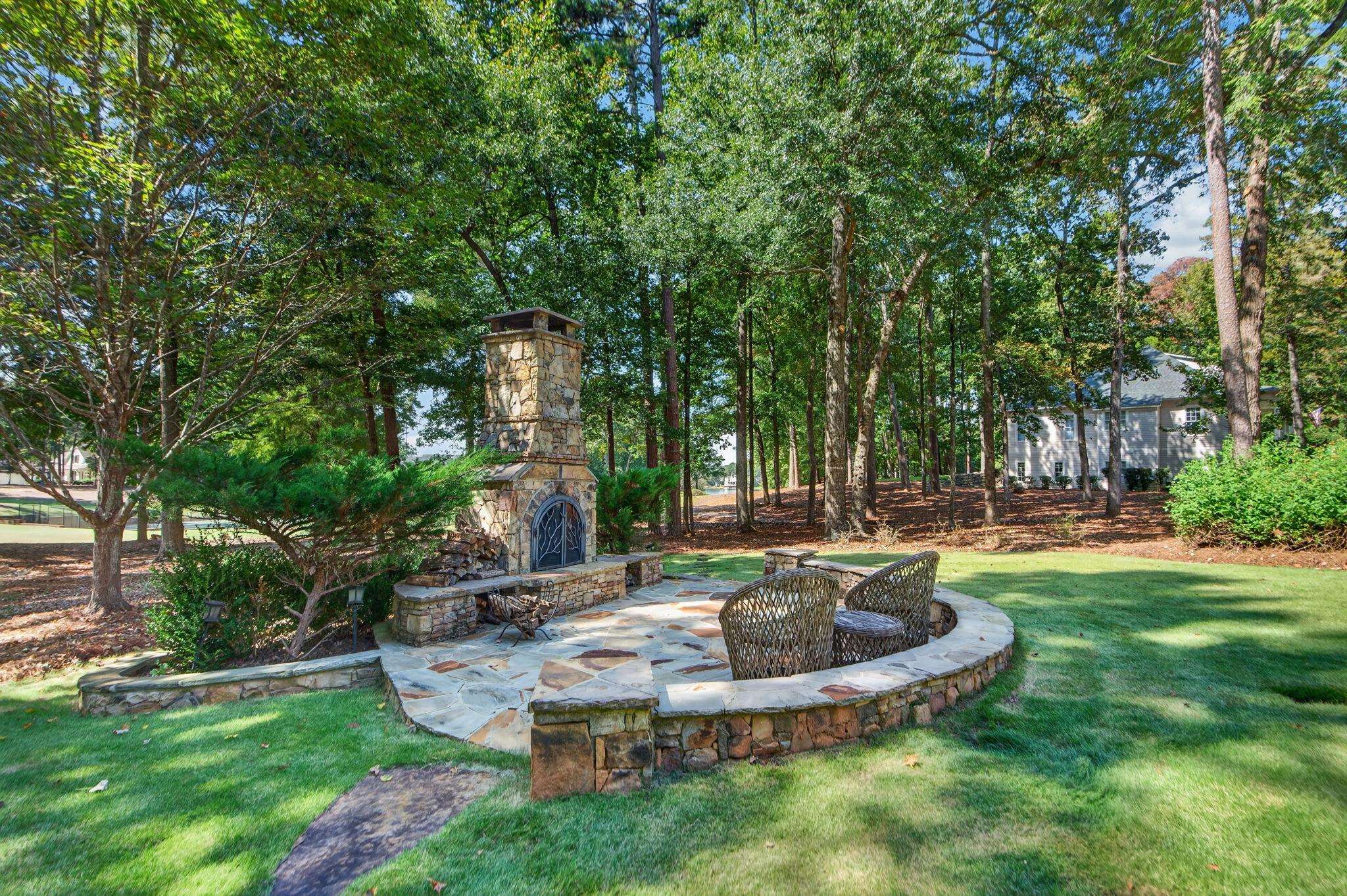 Backyard patio with a stone fireplace, curved seating wall, and woven chairs beneath mature trees.
