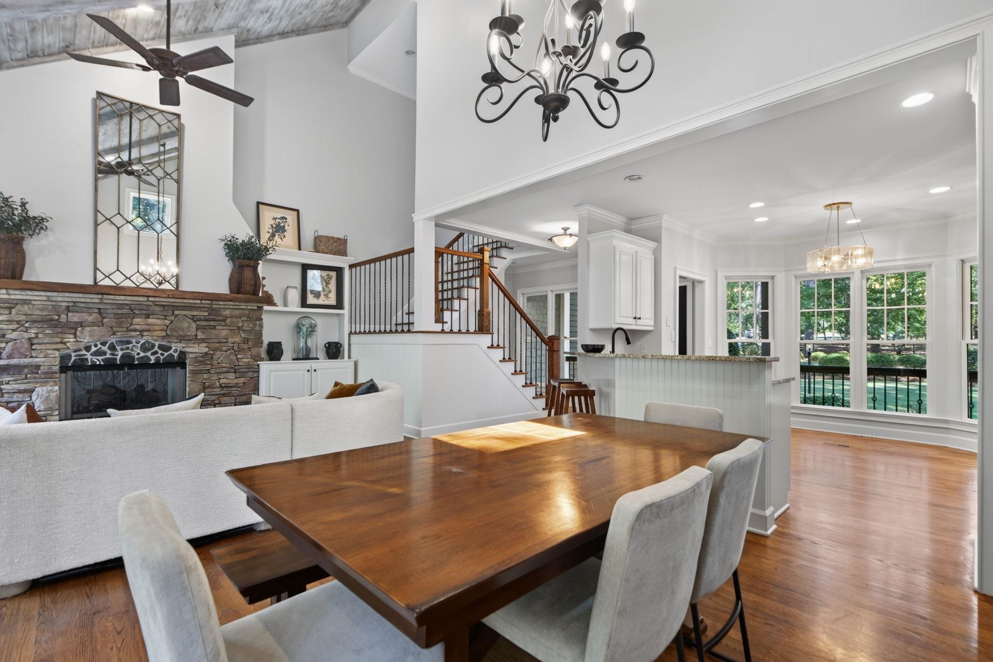Open living and dining area with a stone fireplace, wood table, staircase, and tall windows.